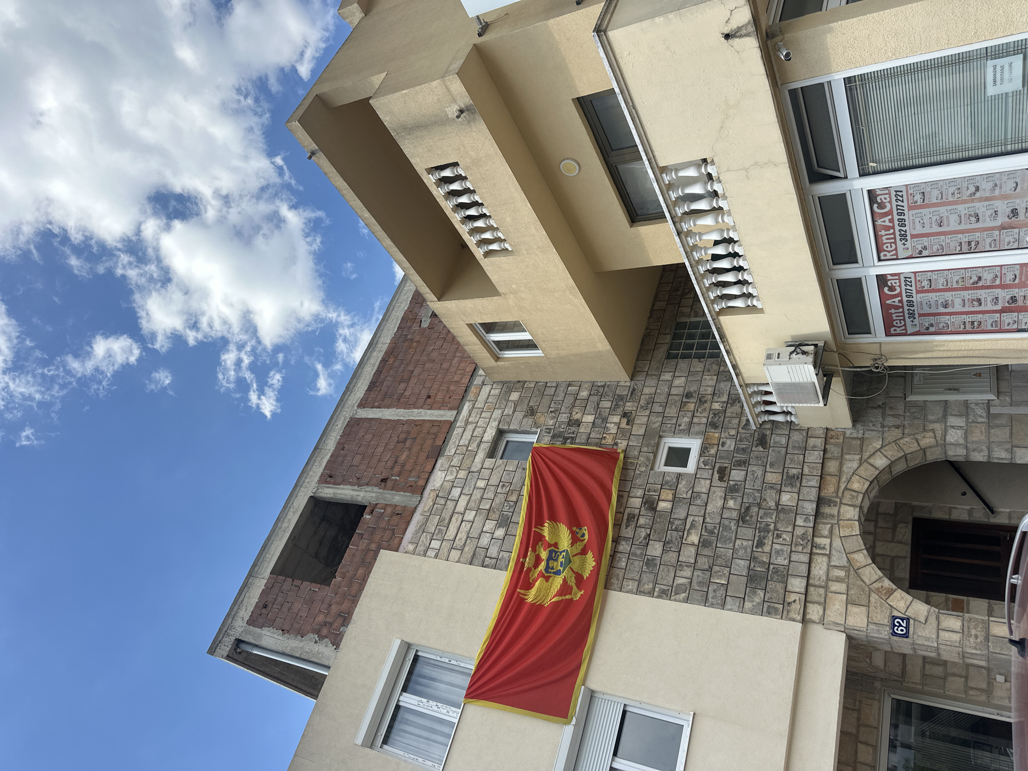 The image shows a multi-story building in Budva, Montenegro, under a blue sky with scattered clouds. The building's facade is a mix of materials, including brick, stone, and painted surfaces. A Montenegrin flag hangs vertically on the building. "Rent A Car" signs are visible on the windows of the building. The number "62" is displayed near an arched entrance.