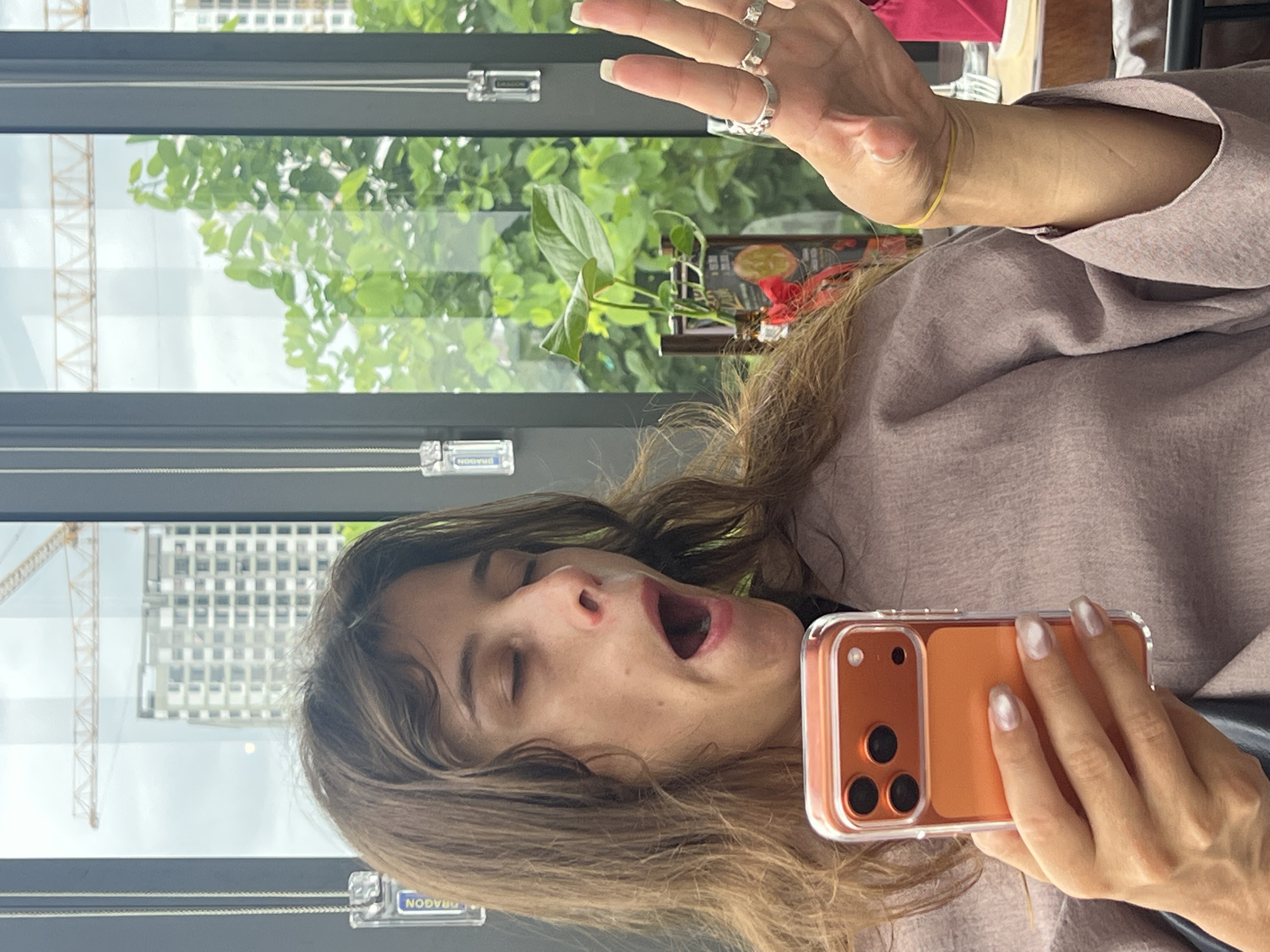 A woman with medium-length brown hair is seen in a close-up shot, yawning with her eyes closed and mouth wide open. She is holding an orange-cased iPhone in her left hand. Her right hand is raised, displaying several rings on her fingers. She is wearing a light purple garment with wide sleeves. In the background, through a window, a construction crane and a tall building are visible, suggesting an urban setting in Đà Nẵng, Vietnam. Lush green trees are also prominent outside the window.