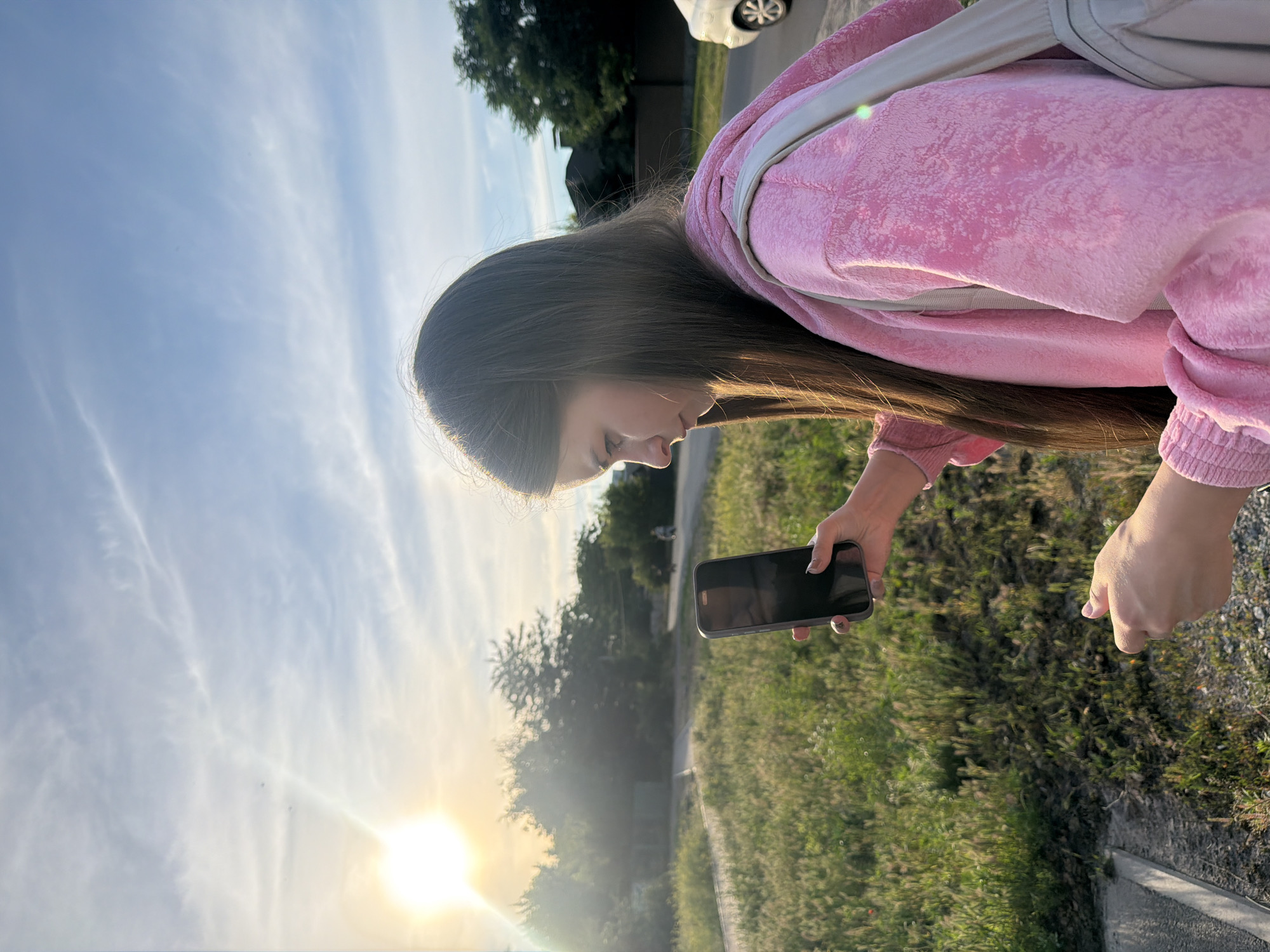 The image shows a young woman with long brown hair wearing a pink hooded jacket. She is outdoors, possibly in a suburban or rural area, with a grassy field and trees visible in the background. The sun is low in the sky, suggesting it is either sunrise or sunset, and casts a warm glow across the scene. The sky is partly cloudy with wispy white clouds. The woman is holding a smartphone in her right hand and appears to be looking at it. Her left hand is extended and pointing downwards. There is a white car visible in the far background. The overall mood is relaxed and casual. The image was taken in Dnipro, Ukraine.