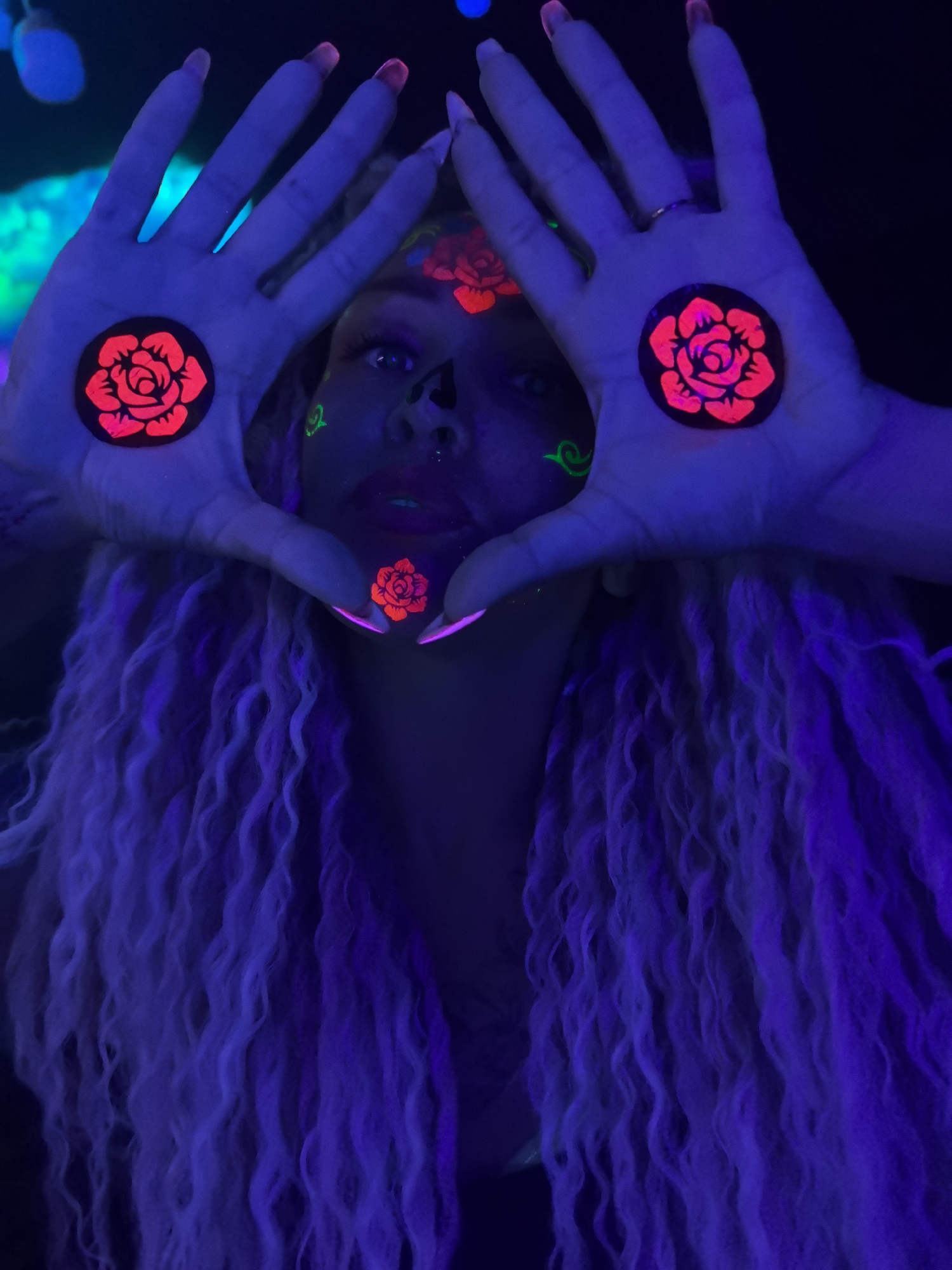 A person with blonde, curly hair is posing in a dark environment illuminated by ultraviolet light. The person has neon face paint and stickers on their forehead, nose, and palms. The stickers are in the shape of roses. The person is holding their hands up, forming a triangular shape around their face. The location is not discernible from the image.
