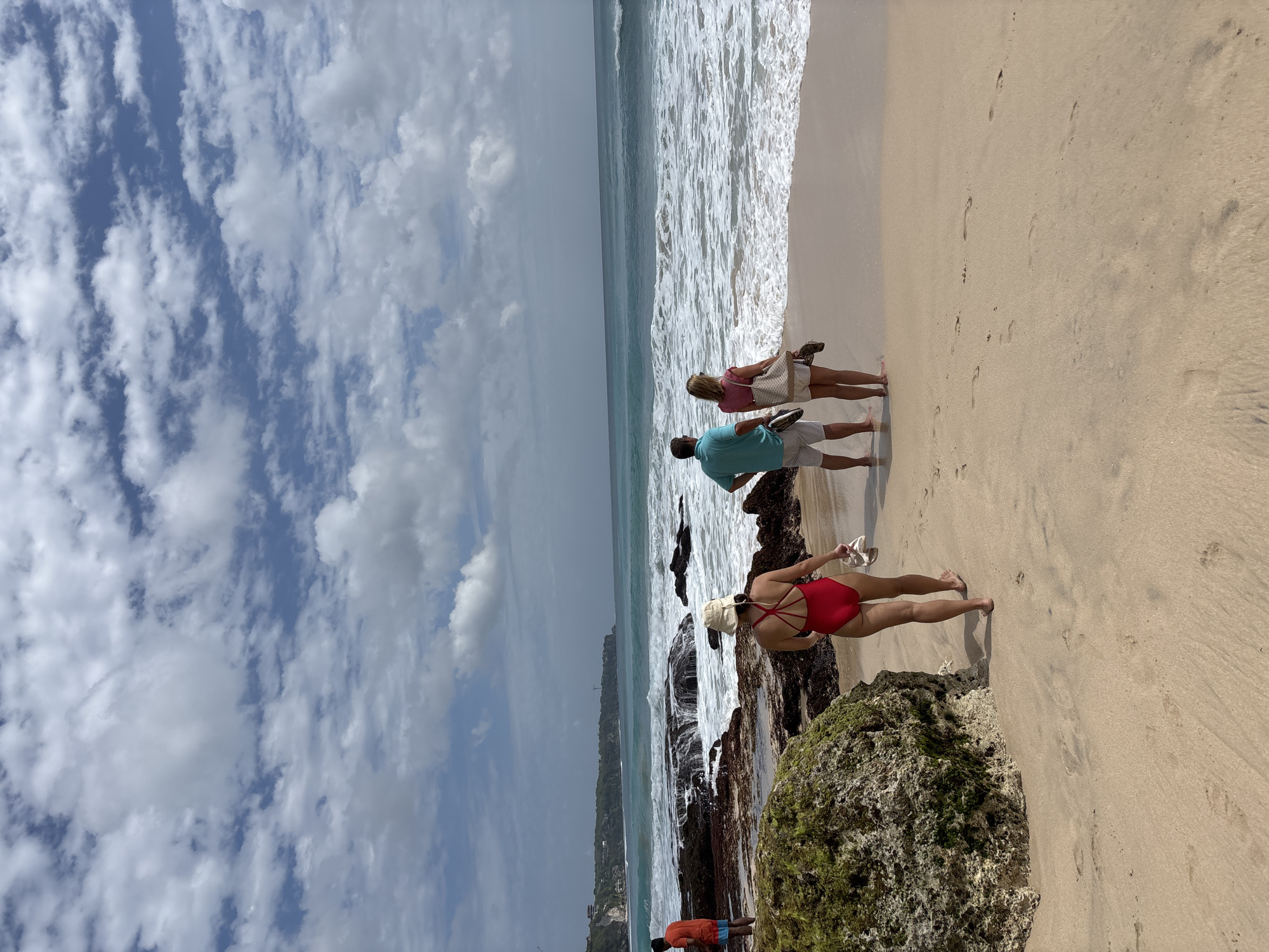 The image depicts a tropical beach scene in Kuta Selatan, Indonesia, under a partly cloudy sky. In the foreground, a person wearing a red swimsuit and a sun hat walks away from the camera on a sandy beach, carrying a pair of sandals. To their right, a couple stands facing the ocean, with the man in a teal polo shirt and white shorts, and the woman in a pink top and white shorts, also holding sandals. Further left, a man in an orange t-shirt and blue shorts stands near some rocky outcrops. The ocean features moderate waves crashing onto the shore, with a rocky coastline visible in the distance. A large, moss-covered boulder is prominent in the left foreground.