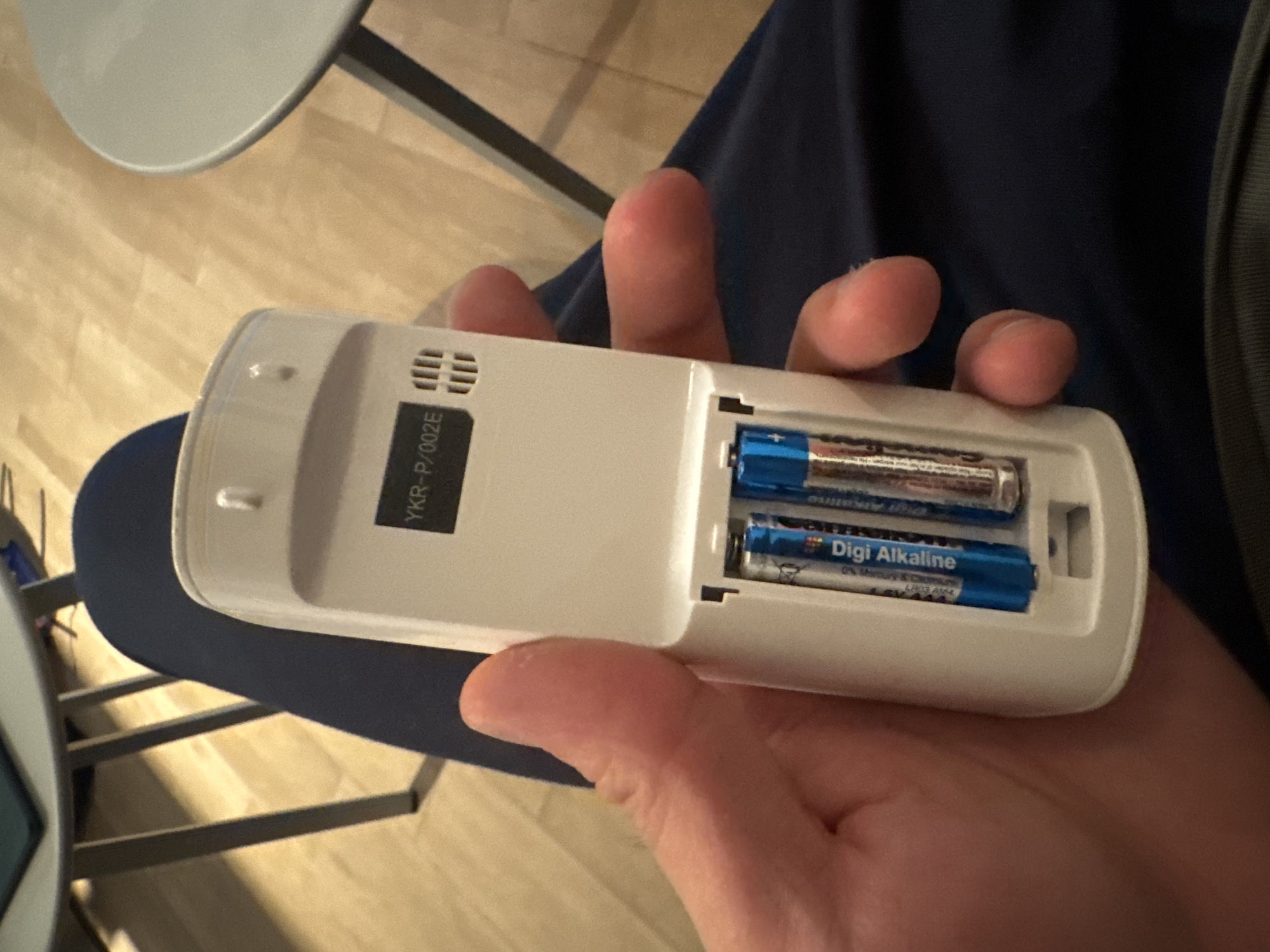 A person's hand is holding a white remote control, with the battery compartment open. Two blue and silver alkaline batteries are visible inside the compartment. The remote has "YKR-P/002E" printed on its back. The scene appears to be indoors, with a wooden floor visible in the background, and parts of a gray table and dark blue fabric. The location is Unknown, Unknown.