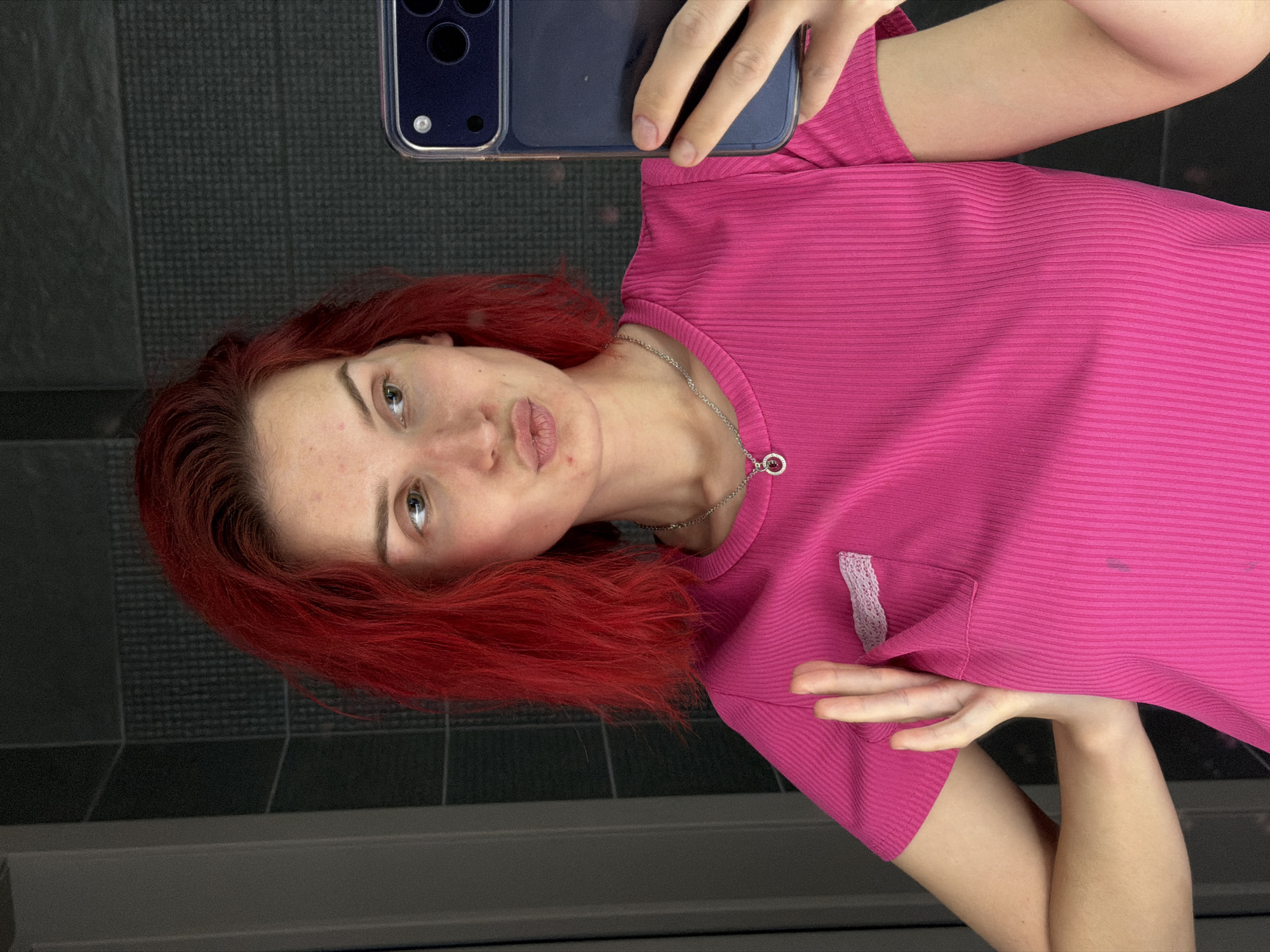 A woman with red hair and a pink shirt is taking a selfie with a phone. She is in a room with gray tiled walls. A location in the city of Chaikovsky, Russia cannot be confirmed.