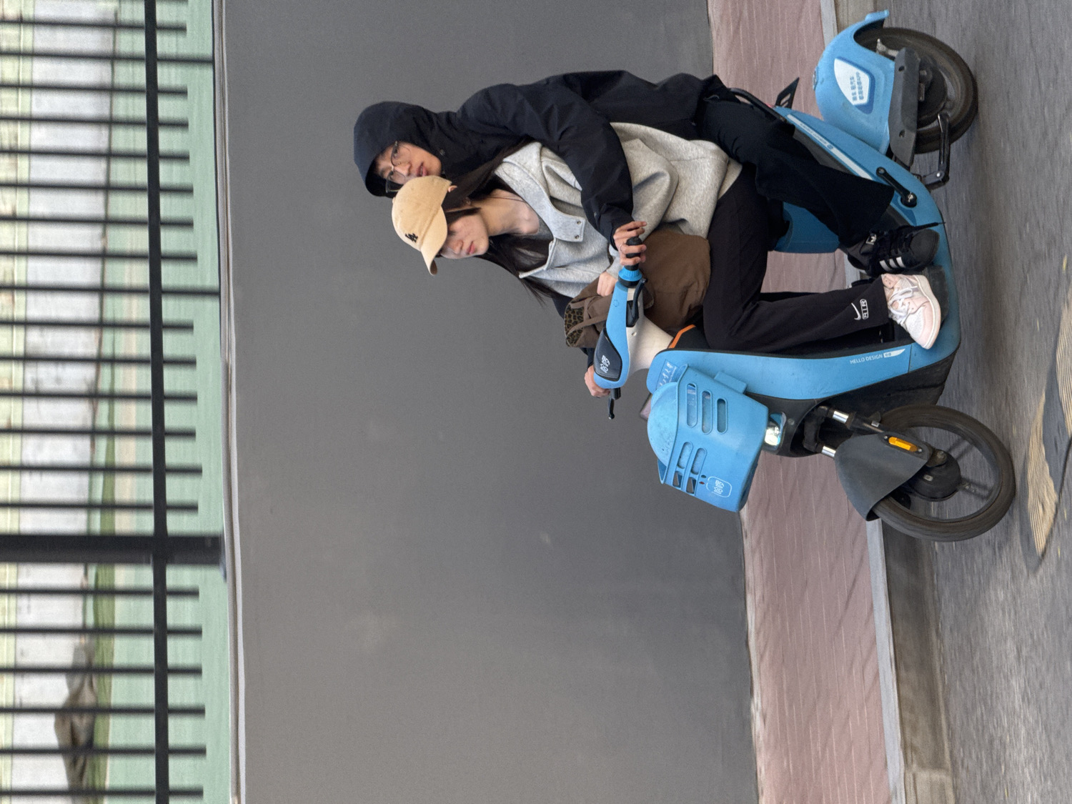 Two young individuals are depicted riding a blue electric scooter outdoors. The image is rotated 90 degrees clockwise, showing them moving along a paved surface.
The person in front, likely driving, wears a beige baseball cap, glasses, a light grey hooded sweatshirt, dark track pants with a white "NIKE" logo on the left leg, and light pink sneakers. They hold the handlebars, and their gaze is directed forward.
Riding as a passenger behind the first person is a second individual, wearing a dark hooded jacket, glasses, dark pants, and black sneakers. They appear to be holding onto the driver.
The scooter itself is bright blue, featuring a front basket area with square cutouts and Chinese characters. On its side, the text "HELLO DESIGN" is visible, along with more Chinese characters. The scooter has black wheels and a front fender.
In the background, to the left (top of the image when rotated correctly), there's a structure with horizontal green slats, possibly a fence or blinds, casting faint reflections. To the right (bottom of the image when rotated correctly), a plain grey wall or surface is visible, transitioning to a light brown/pinkish paved area. The setting appears to be a street or pathway. The lighting suggests it is daytime, possibly overcast.
The activity is the two people riding the scooter, seemingly focused on their journey. The location is contextualized as Zhongtun, China.