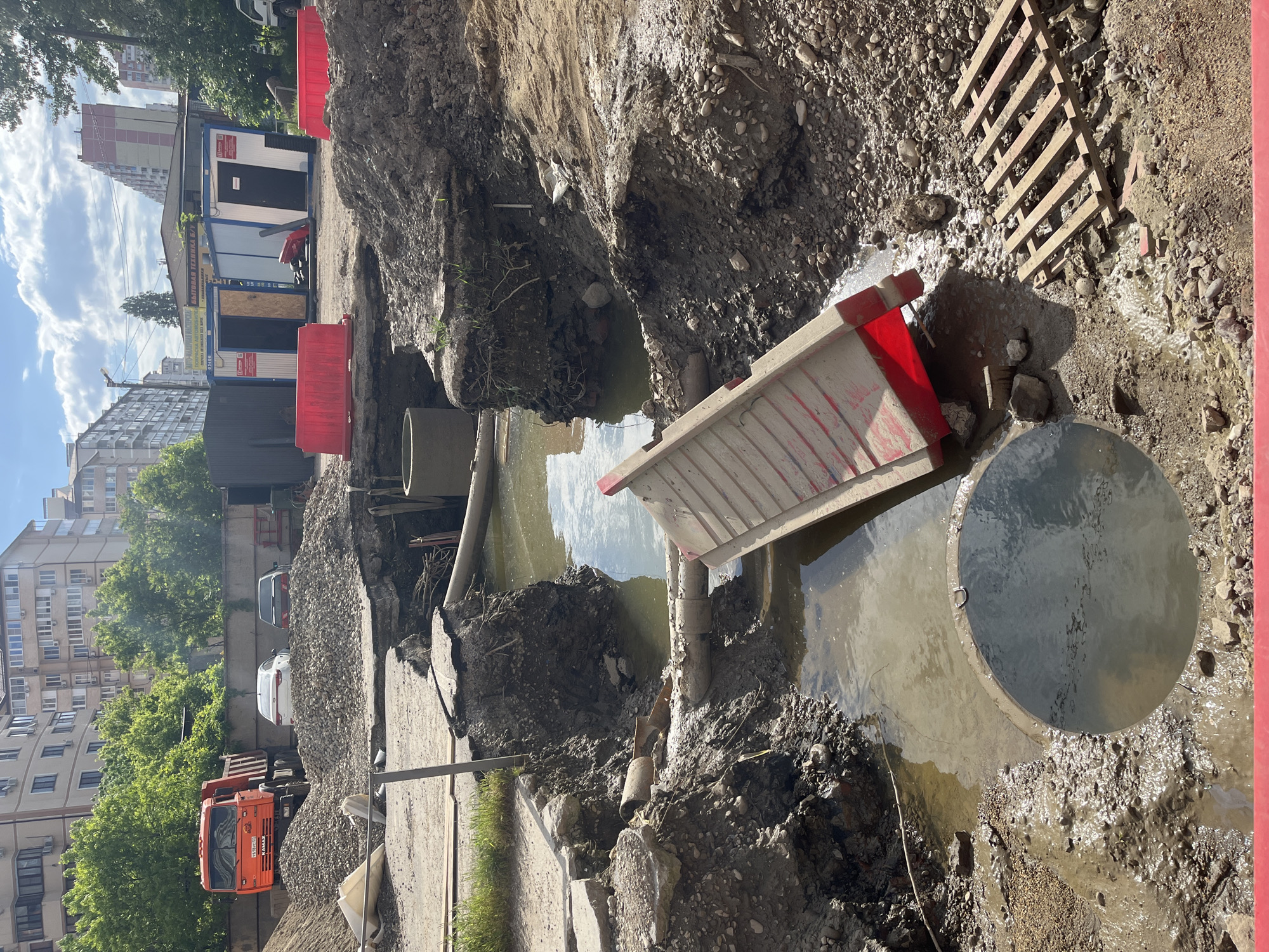 The image depicts a construction site in an urban area, likely Kozet, Russia, given the truck with a Russian license plate visible in the background. A large excavation has been dug, filled with muddy water, and littered with construction materials. Visible in the water are sections of concrete pipe, a fallen barrier with red accents, and a round metal manhole cover partially submerged. The edges of the excavation are lined with dirt and stones. In the background, there are residential buildings, trees, parked cars (white and gray), and construction trailers, suggesting the site is located within a populated area. An orange truck is visible on the left, possibly involved in the construction work. A wooden pallet is visible on the ground surface next to the construction. Red safety barriers are set along the excavation for hazard protection. The scene is captured during daylight, under a partly cloudy sky.