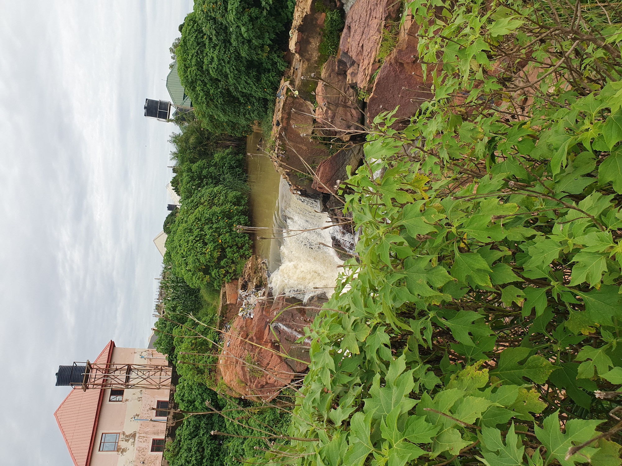 The image depicts a waterfall in Gura Top, Nigeria, with lush green foliage in the foreground and midground. A light pink building with a red roof and a tall water tank stands to the left, overlooking the cascading water. The water is a muddy brown, suggesting it is carrying sediment. Several large rocks, varying in color from reddish-brown to grey, are visible within and around the waterfall. The sky is overcast with grey clouds, indicating a potentially humid or rainy day. Scattered debris is visible on the rocks near the top of the waterfall. Further in the background, more buildings and dense vegetation can be seen, with additional water tanks positioned on some rooftops. The overall scene conveys a sense of a natural landscape integrated with human development.