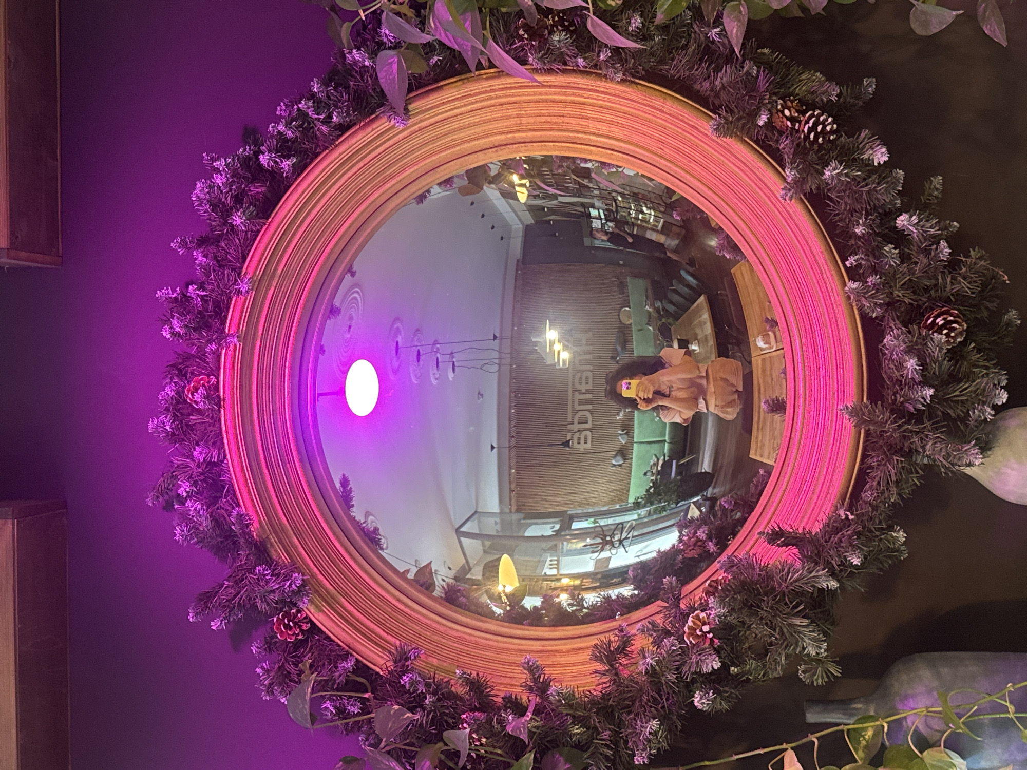 A woman holds a smartphone, taking a selfie in a large, ornate, circular mirror. The mirror is framed with garland, including pine branches and pinecones, illuminated by pink and purple lights. The reflection in the mirror shows a cafe interior with wooden accents, green seating, and tables. The woman is wearing a light-colored outfit. The overall impression is a festive and cozy atmosphere, likely in a cafe in Saint Petersburg, Russia.