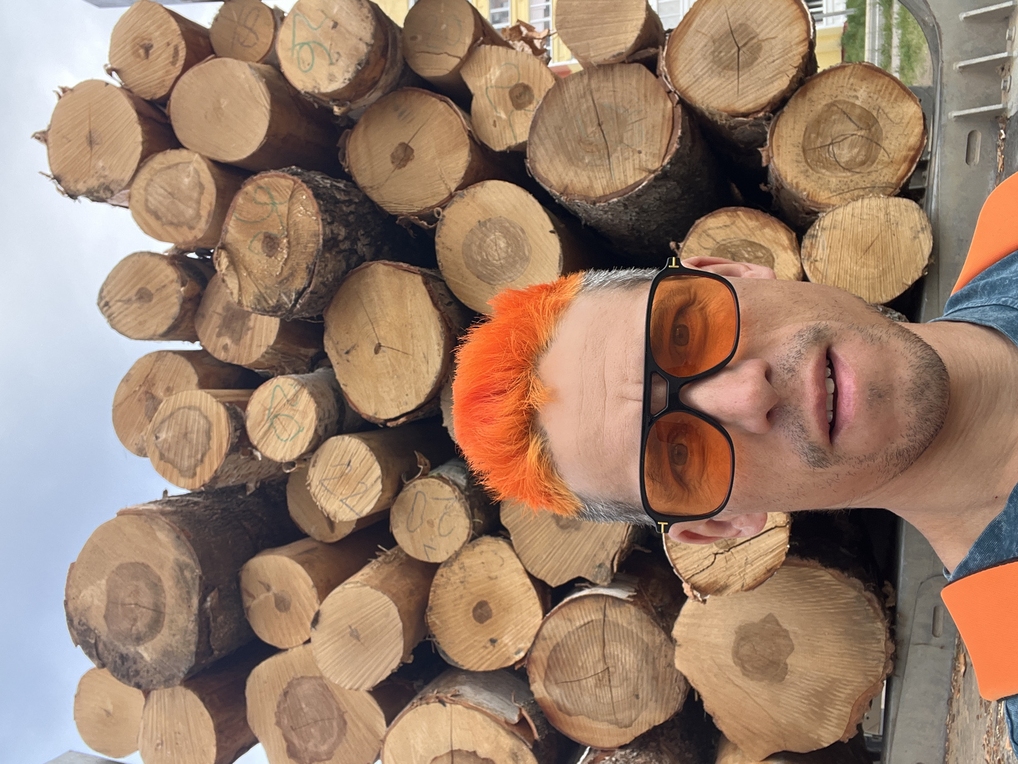 A man with orange hair and dark-rimmed glasses is positioned in front of a large stack of cut logs. The logs are densely packed and bear markings on their ends. The man is wearing a denim shirt with an orange sleeve. The background includes a glimpse of a building with yellow and white walls and a cloudy sky. The location is Chaikovsky, Russia.
