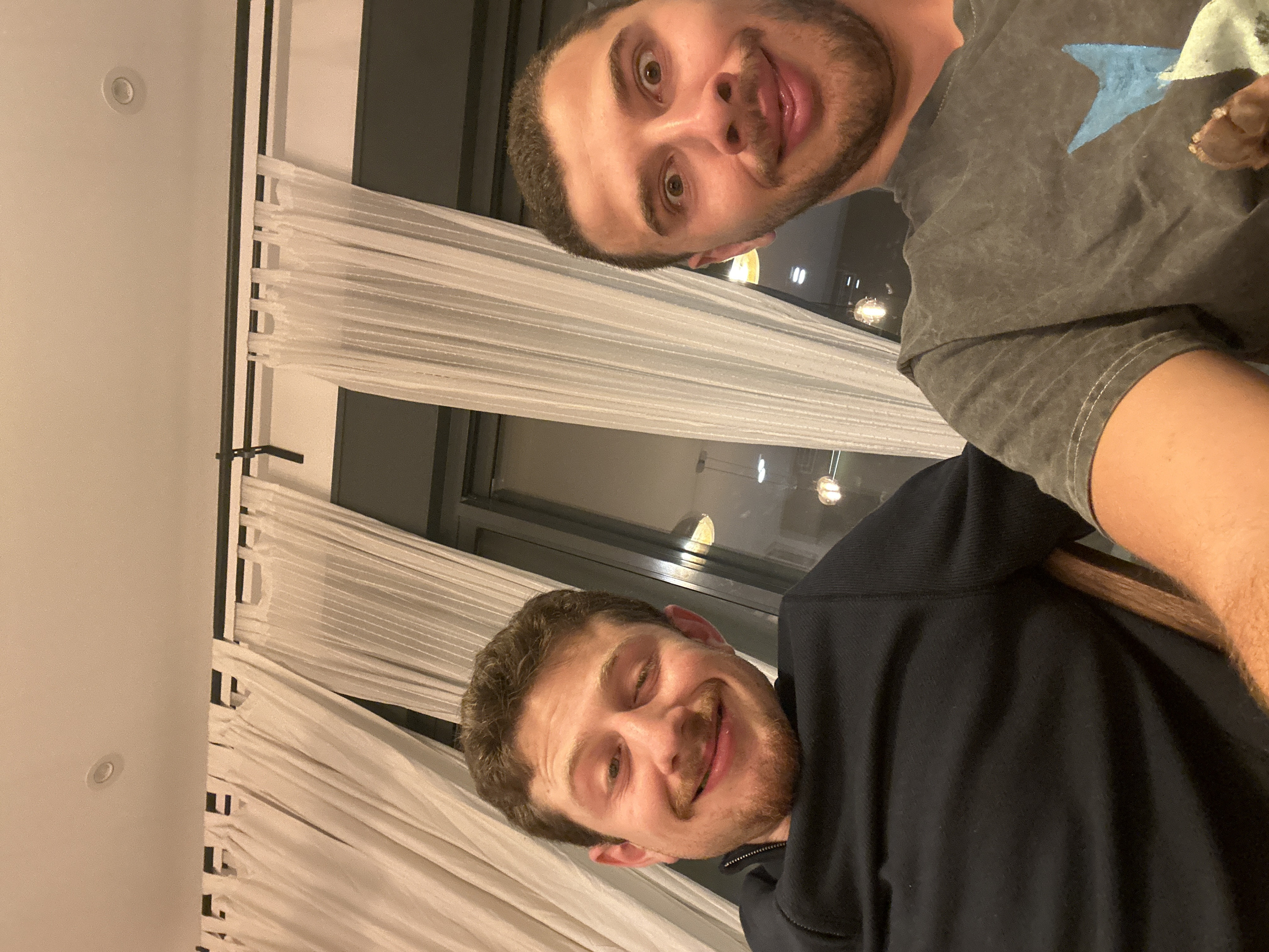 Two men pose for a selfie indoors at night. One man, with dark hair and a goatee, wears a grey t-shirt with a blue star. The other man, with light brown hair and a mustache, wears a black zip-up jacket. White curtains hang in the background by a window reflecting lights.
