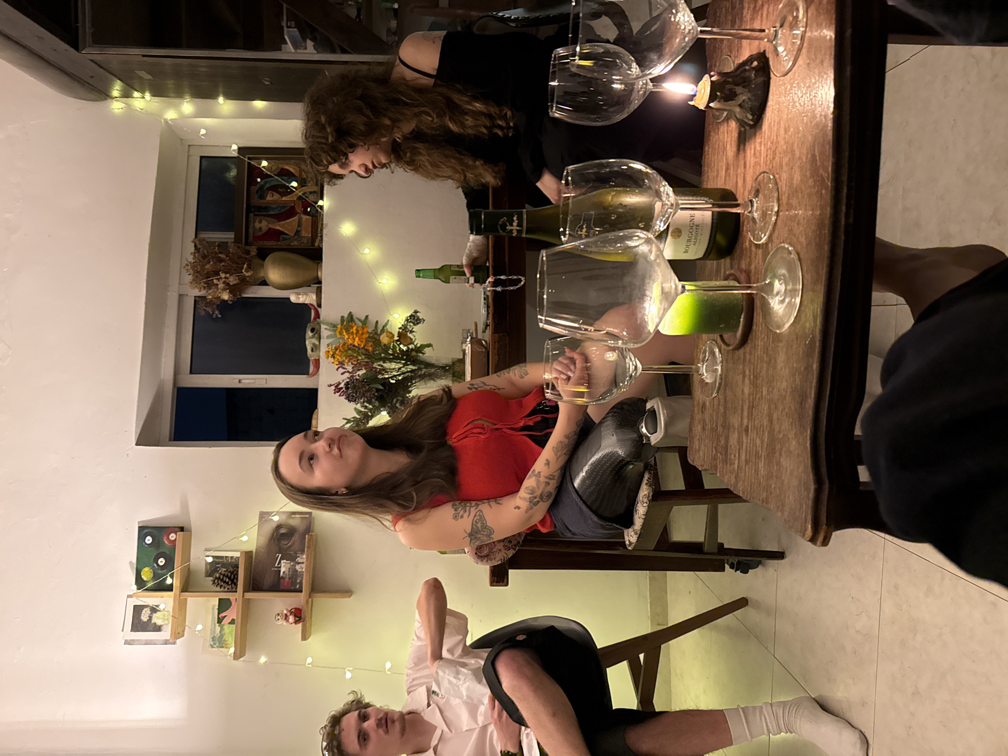 The image captures an indoor social gathering, seemingly a dinner party, with four individuals visible. A woman with long, curly brown hair, dressed in black, stands at a wooden table laden with wine glasses, bottles of wine (including Bourgogne Aligoté), and a lit candle. Another woman, with brown hair and tattoos, wears a red top and sits at the table, appearing to engage in conversation. A person wearing a prosthetic leg is seated next to her. In the foreground, a man wearing a white shirt and dark shorts sits on a chair, also looking engaged in conversation. The fourth individual is partially visible on the right, wearing a black shirt. The room is illuminated with string lights. A window and a bookshelf with various items are visible in the background. The floor appears to be tiled. The location is assumed to be an apartment in Tel-Aviv, Israel. There are no visible signs that confirm this location, but it is mentioned in the prompt. The people are drinking wine and conversing.