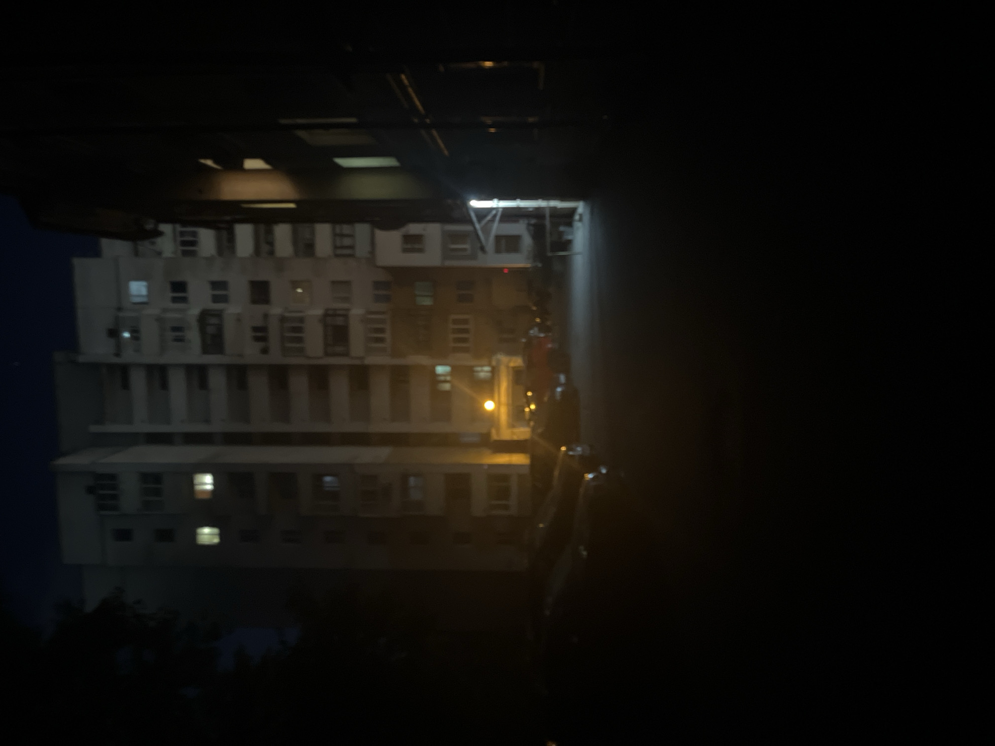 A nighttime, slightly hazy, medium-shot captures a street in a city. In the background, a tall, multi-story apartment building is visible, with some windows illuminated by artificial light. To the right, another building, possibly commercial, is partially visible, also with some interior lighting. In the foreground, several cars are parked on the asphalt. The overall atmosphere is dark and quiet, typical of a residential area at night.