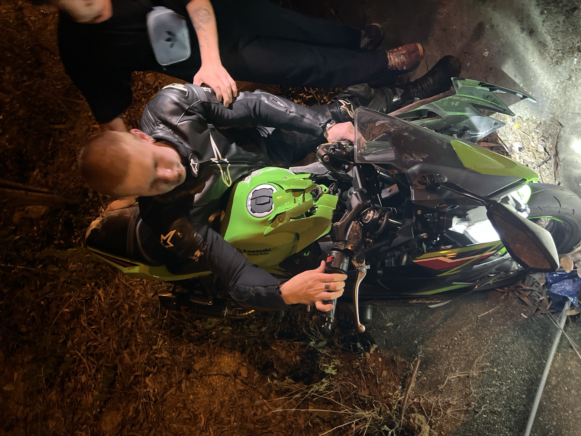 A man in black leather motorcycle gear lies next to a green and black Kawasaki motorcycle that appears to be wrecked. The motorcycle is laying on its side with damage to the front fairing. Another man, also wearing black, kneels next to the man and motorcycle, seemingly attending to the situation. The location is a dark outdoor area with ground covered in dirt, leaves, and debris. A light source illuminates the scene, possibly from a vehicle or street light. Based on the context provided (Bac Natural Heritage Area Trang, Vietnam), this scene could potentially be located somewhere within or near this area.