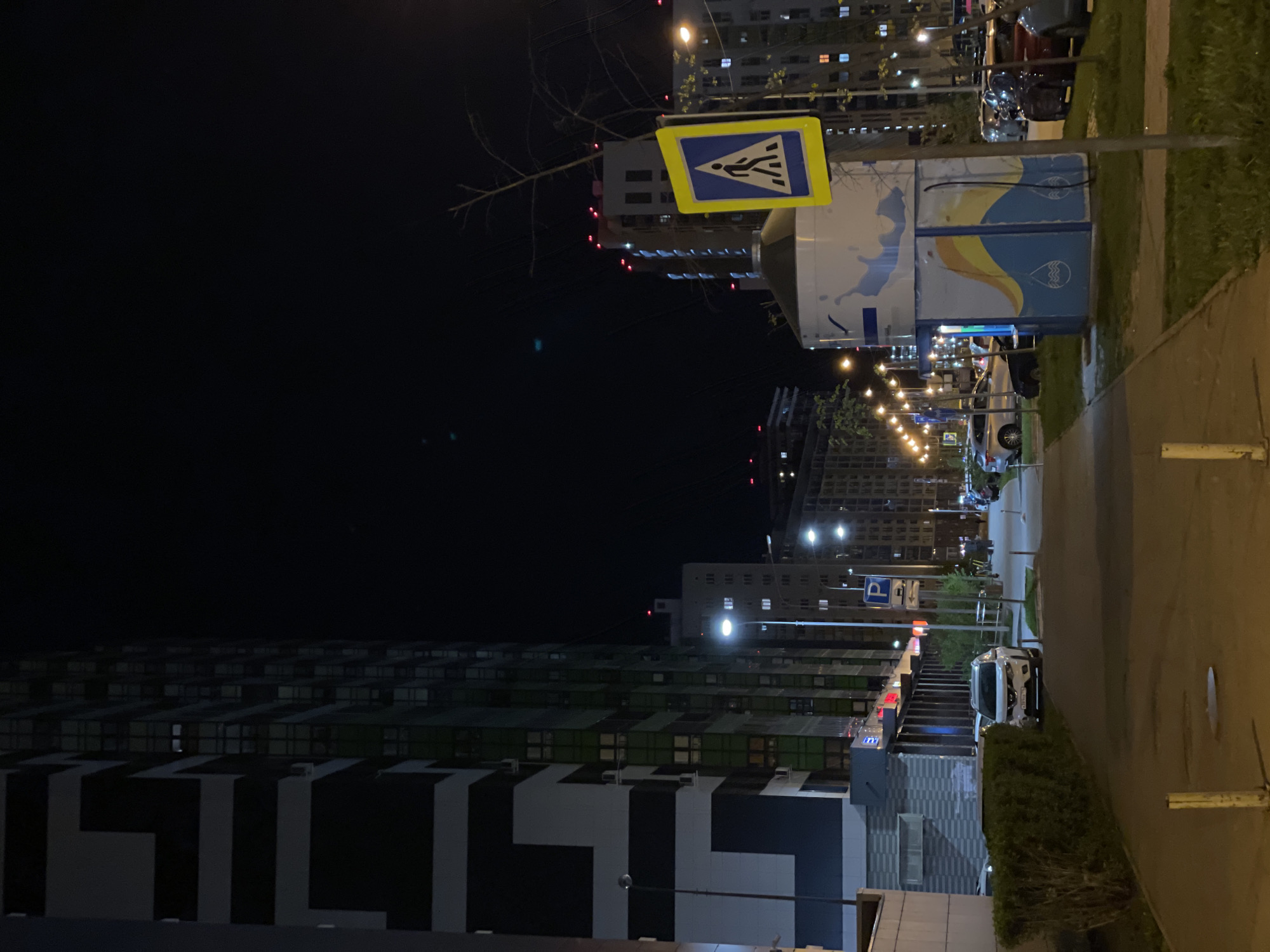 The image shows a night scene in Kazan, Russia, along a sidewalk. The street is lined with modern high-rise apartment buildings with illuminated windows. A pedestrian crossing sign is visible near a sidewalk vending stall or kiosk. Streetlights provide illumination. The area appears to be a residential neighborhood. There are also cars in the scene and greenery along the sidewalk.