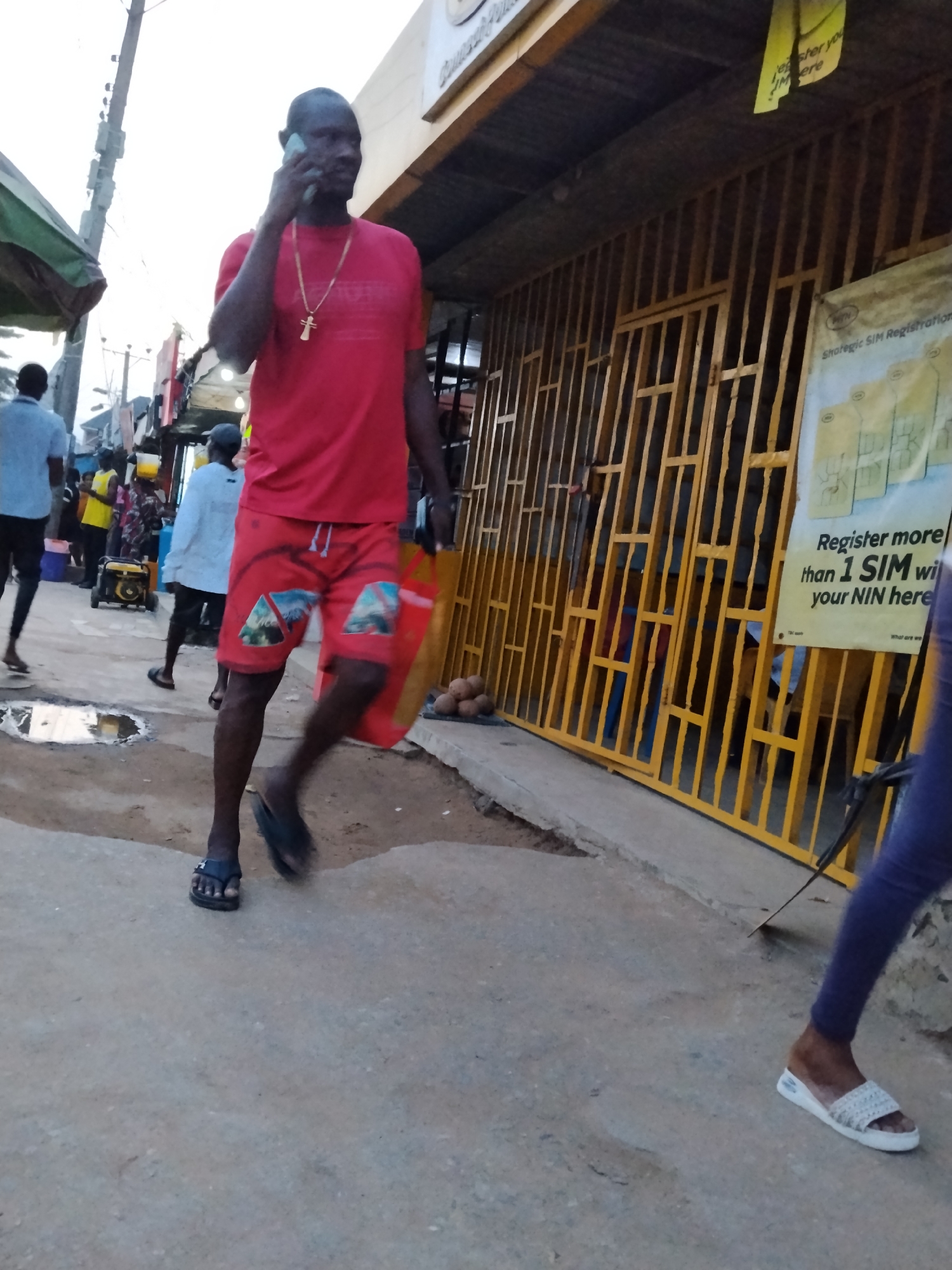 A man wearing a red shirt, red shorts, and sandals is walking down a street in Ura Nta Umuarandu, Nigeria while talking on a cell phone. He is carrying a red shopping bag. He has a gold cross necklace on. In the background, a building has a yellow, barred storefront. A sign for SIM registration is posted on the building. Other people are walking down the street. The street is paved, and there is a puddle of water.