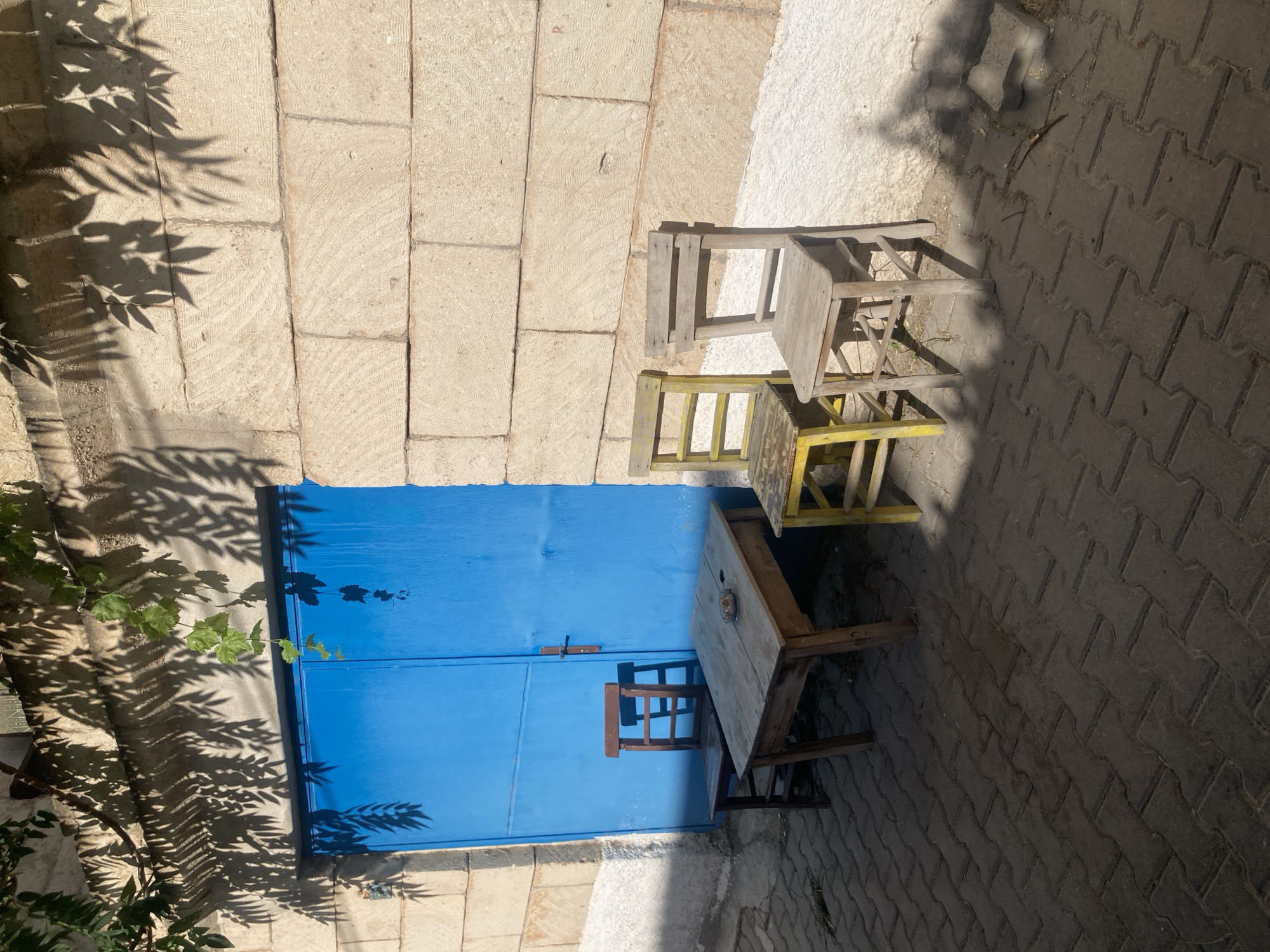 A sunlit exterior scene in Avanos İlçe Merkezi, Turkey, features a bright blue double door set into a wall constructed of large, light-colored, roughly hewn stones. Overhanging the top of the wall are branches with green leaves, casting dappled shadows on the stone. In front of the door, on a paved patio made of interlocking dark grey pavers, are a simple wooden table and three chairs. The table is made of light-colored wood with visible grain and has a small object resembling an ashtray or dish on its surface. To the left of the table is a dark wooden chair with a slatted back. In front of the table and slightly to the right is a small, brightly painted yellow chair. To the right of the table and yellow chair is a weathered, light-colored wooden chair with a horizontal backrest. The ground is partially covered in shadows from the leaves and objects. The overall impression is one of a simple, outdoor seating area.