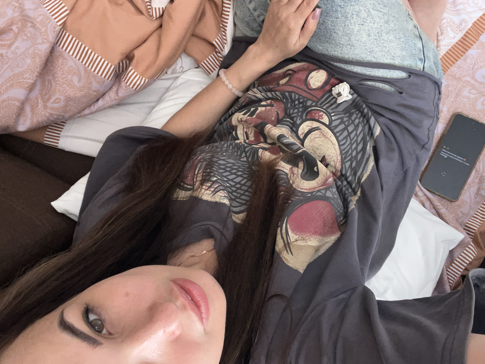 A young woman with long, dark hair is lying down, taking a selfie. She is wearing a grey t-shirt with a graphic print and denim shorts. A light pink beaded bracelet is visible on her wrist. She is on a bed with patterned pillows and blankets. A smartphone is placed on the bed next to her. The scene appears to be indoors, possibly in a bedroom.