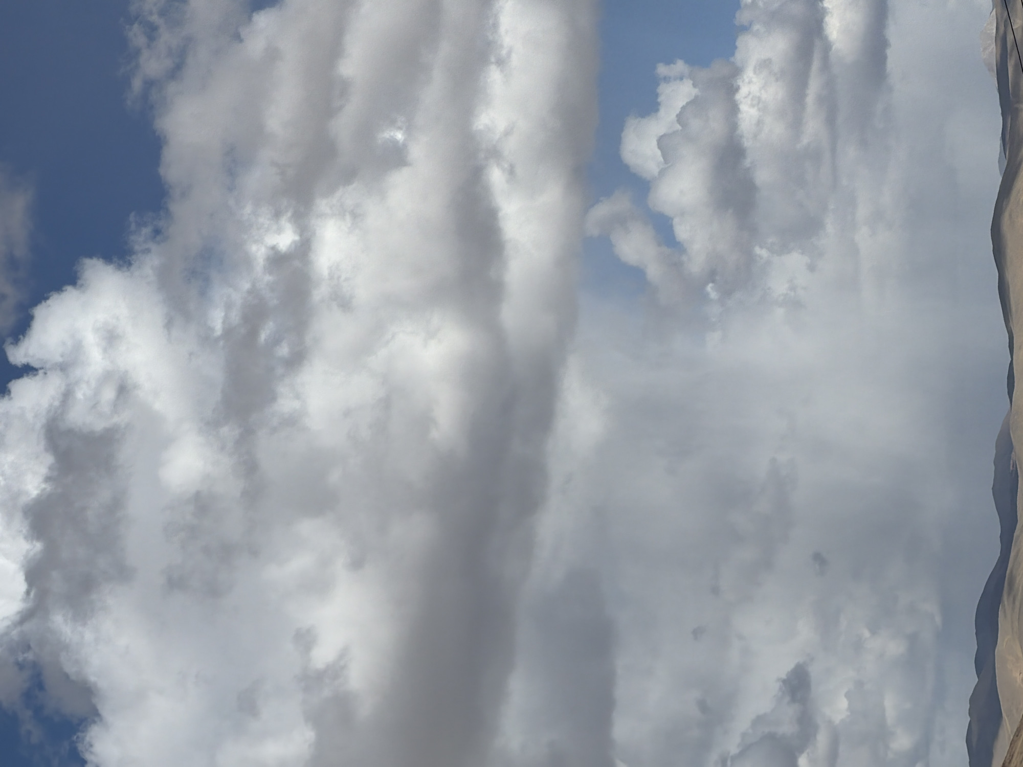 The image displays an expansive outdoor scene dominated by the sky, filled with a diverse array of clouds. When viewed in its correct landscape orientation, the top portion features large patches of clear blue sky, while the majority of the frame is covered by thick, cumulus, and stratocumulus clouds. These clouds exhibit a range of tones, from brilliant white where sunlight hits them directly, to various shades of grey indicating greater density and shadow.
Towards the right edge of the image, a segment of a mountain range or large hills is visible. These geological formations appear rugged and are characterized by light brown and beige hues, suggesting an arid or high-altitude environment. The line where the mountains meet the sky is relatively sharp, indicating clear visibility despite the cloud cover.
The scene captures a daytime moment, evident from the natural light illuminating the clouds and the blue sky. The weather appears dynamic, with both clear sky and substantial cloud formations present. There are no human subjects, man-made objects, or discernible activities taking place. The overall impression is one of natural grandeur and a vast, open landscape, aligning with the mountainous region of Labrang township in China. No visible text is present in the image.