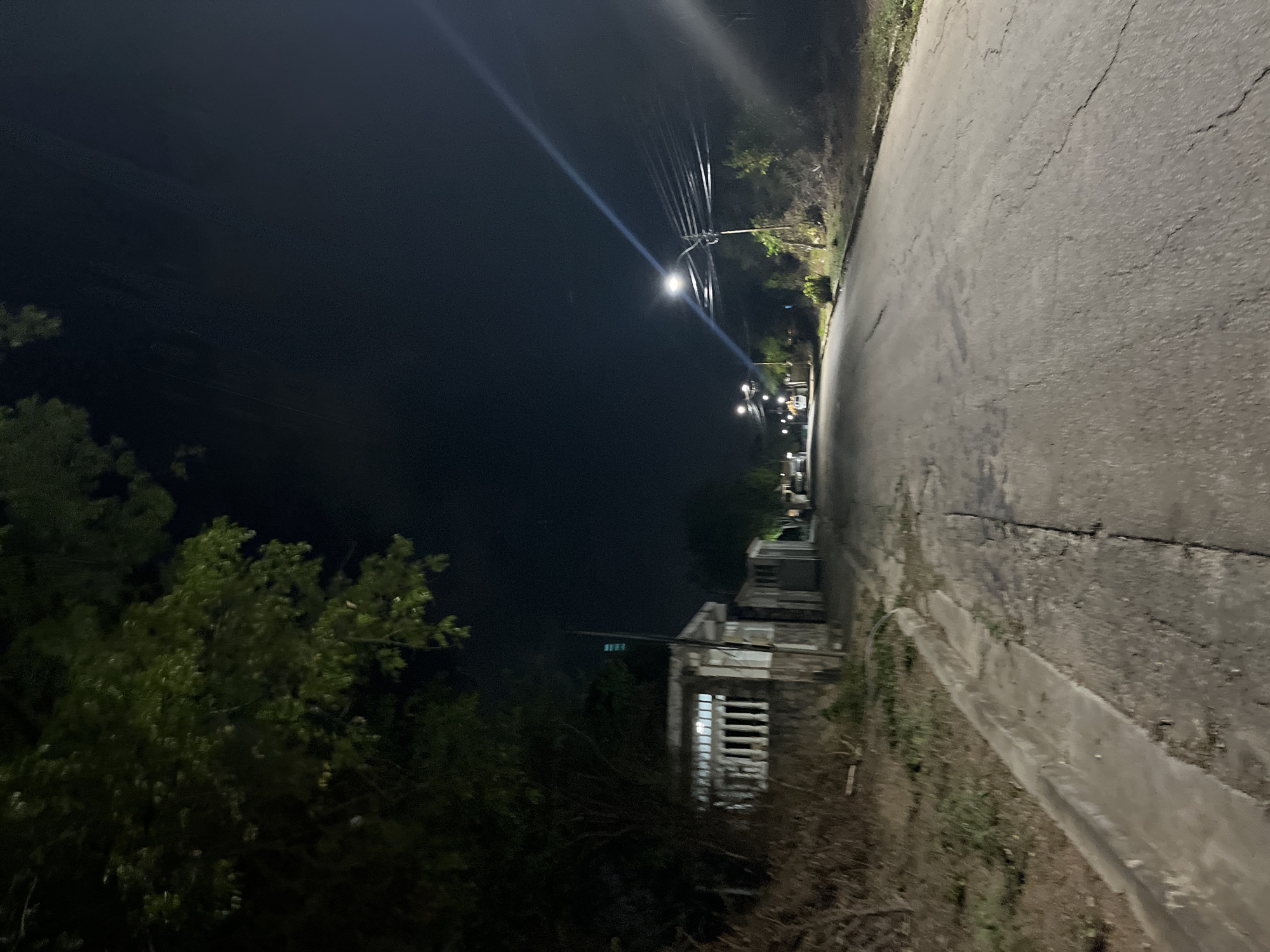The image depicts a dimly lit, paved road at night, likely in a suburban or rural area of Santa Ana, Venezuela. The road is cracked and appears to be in fair condition. To the left of the road, there is a concrete curb and a sloping, unkempt embankment covered with dry brush and some sparse green vegetation. Further up the embankment, a low-lying building with white-barred windows and a stone-like lower section is visible, partially obscured by dark foliage. A vertical pole with a light fixture on top stands near this building.
On the right side of the road, the terrain is also unkempt with dry vegetation and some scattered trees. Several utility poles with numerous wires are visible, illuminated by streetlights, one of which is particularly bright and casts a strong beam across the scene. In the distance, more streetlights are visible along the road, suggesting it is a residential or access road. The sky is dark, indicating nighttime. No people are present in the image. The overall impression is one of quiet, residential nighttime scenery in a Venezuelan town.