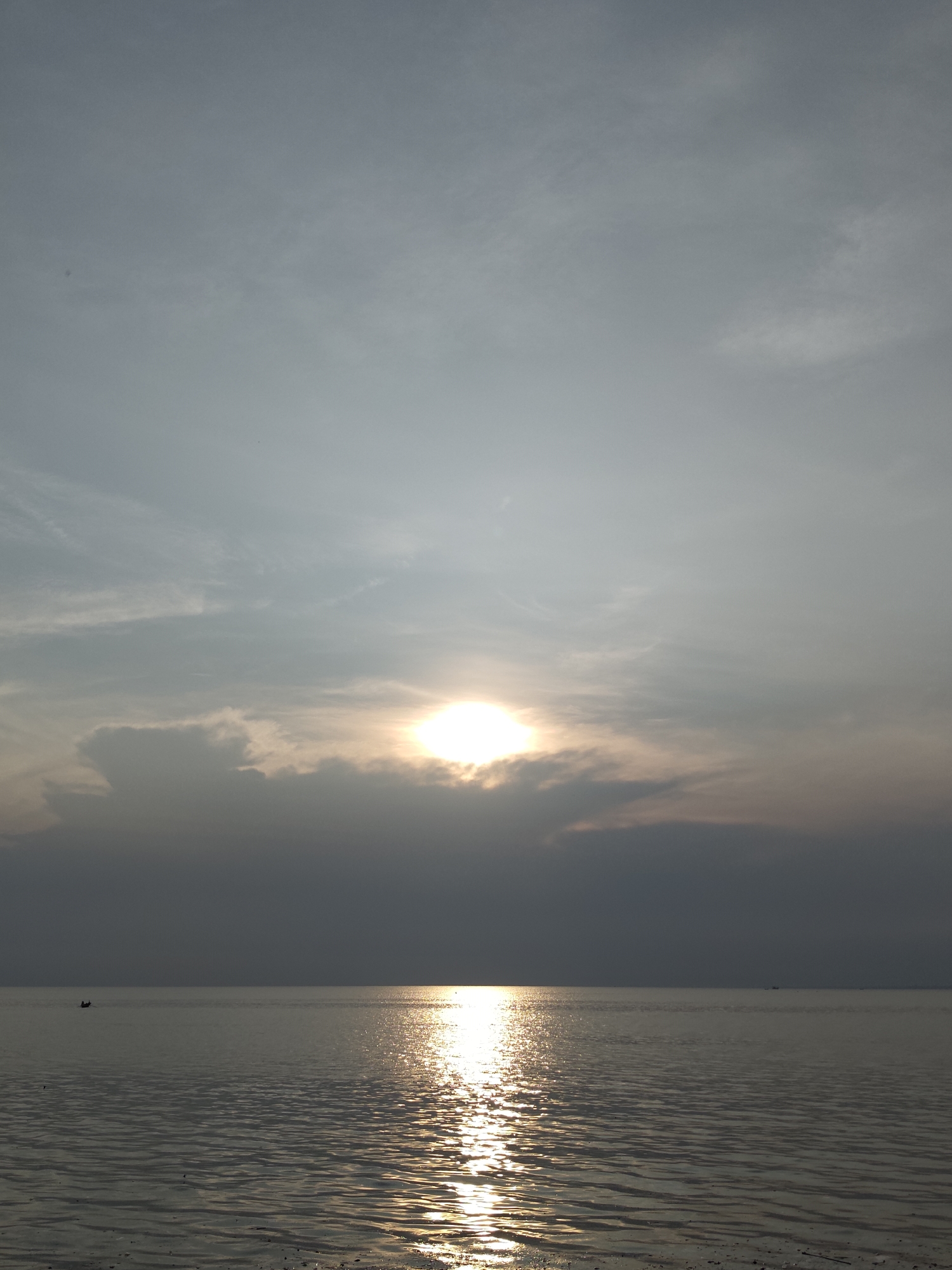 The image shows a seascape under a cloudy sky. The sun is visible, partially obscured by clouds, casting a bright reflection on the water. The water surface is rippled. A small boat is visible on the horizon, left side of the frame. The location is Lhokseumawe, Indonesia.