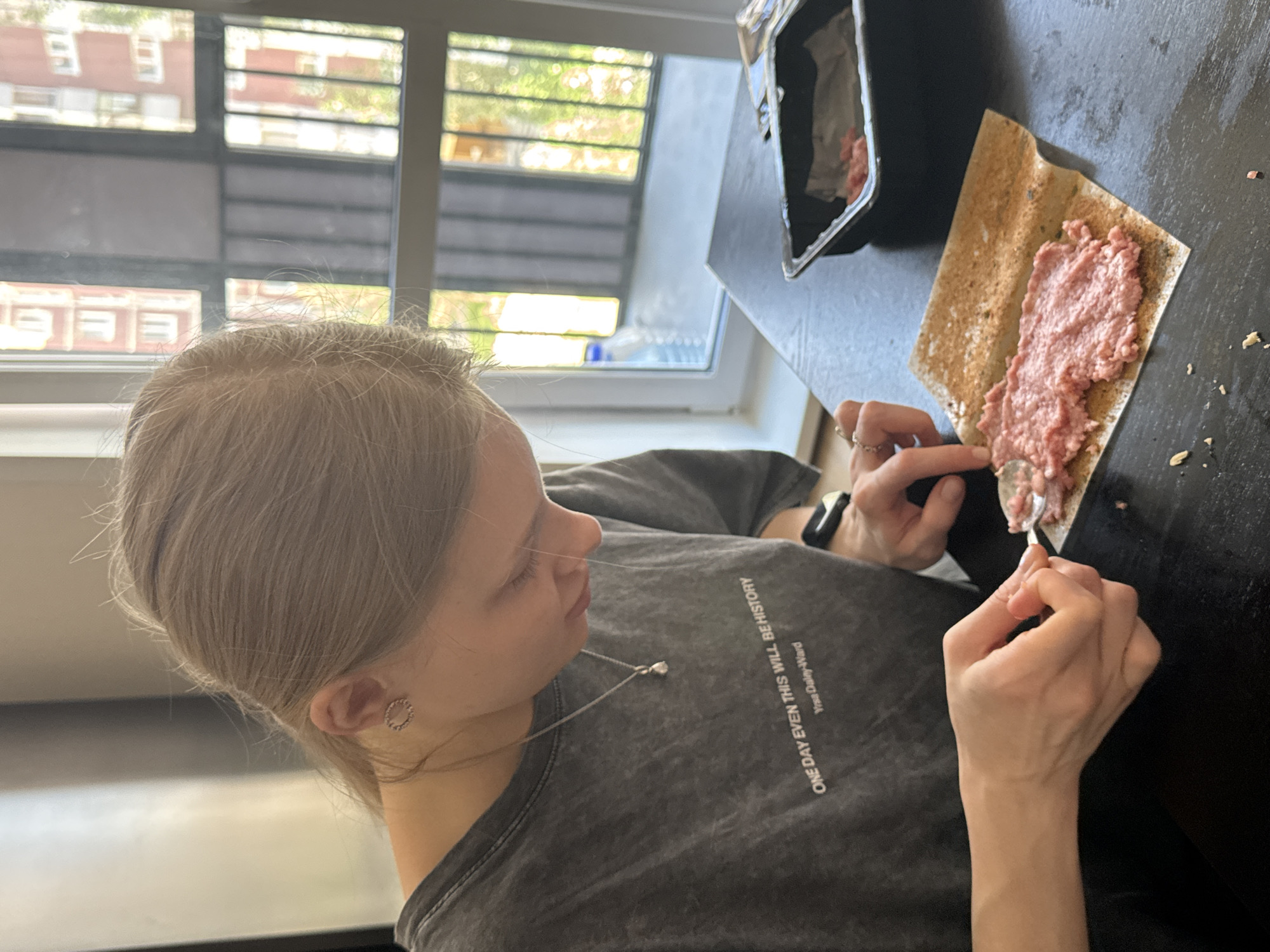 A young woman with fair hair is shown in a medium shot, angled from above and to her left. She is wearing a grey t-shirt with the words "ONE DAY EVEN THIS WILL BE HISTORY Yrsa Daley-Ward" printed on it, and a silver necklace with a heart pendant. In her right hand, she holds a spoon and spreads a pink substance onto a rectangular cracker. The cracker rests on a black tabletop. To the left of the cracker is a small black tray, also containing the pink substance. Background details visible through a window include buildings, suggesting an urban environment. While the city of Perm, Russia is requested in the prompt, there is no supporting information in the image to support that this scene is taking place there.