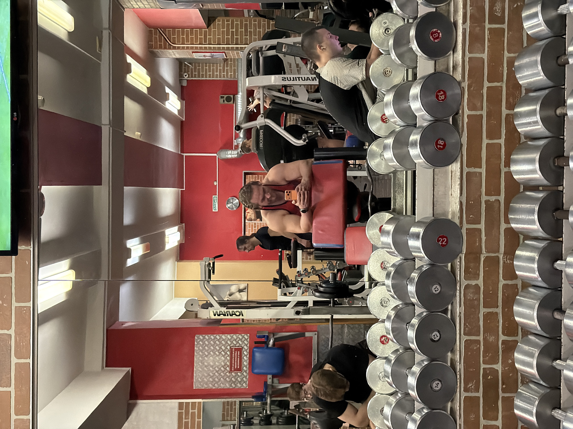 The image depicts the interior of a fitness center, assumed to be located in Saint Petersburg, Russia, based on contextual information. The foreground is dominated by a two-tiered rack of numerous silver dumbbells with red numerical weight markings, ranging from 8 to 22 kilograms. A low, brick-patterned wall serves as the base for the dumbbell rack.
The background features a large mirror reflecting the gym's interior, which has predominantly red walls, interspersed with areas of faux-brick and a silver diamond-plate panel. The ceiling is white with multiple rectangular fluorescent light fixtures and sections painted red. A black LG television screen is mounted high on the left. A Russian "ПОЖАР" (FIRE) sign with an arrow is visible on a red wall in the upper left quadrant.
Five individuals are present. In the mid-ground, a man with dark hair, wearing a black t-shirt, is leaning forward over a piece of equipment, with his face obscured. To his right, a muscular man with medium-length dark hair, wearing a red tank top, leans on a red-padded bench while engrossed in an orange smartphone held in both hands. In the mirror's reflection, to the right, a young man with short dark hair, wearing a black and white striped t-shirt, is seated, looking down at a brightly lit smartphone. Further left in the reflection, another man with dark hair tied back, wearing a black t-shirt, stands near a multi-gym machine. The back of a fifth person, wearing a black top, is visible in the reflection, positioned on a piece of exercise equipment. The scene shows individuals engaged in personal activities within a gym setting, primarily resting or using mobile devices.