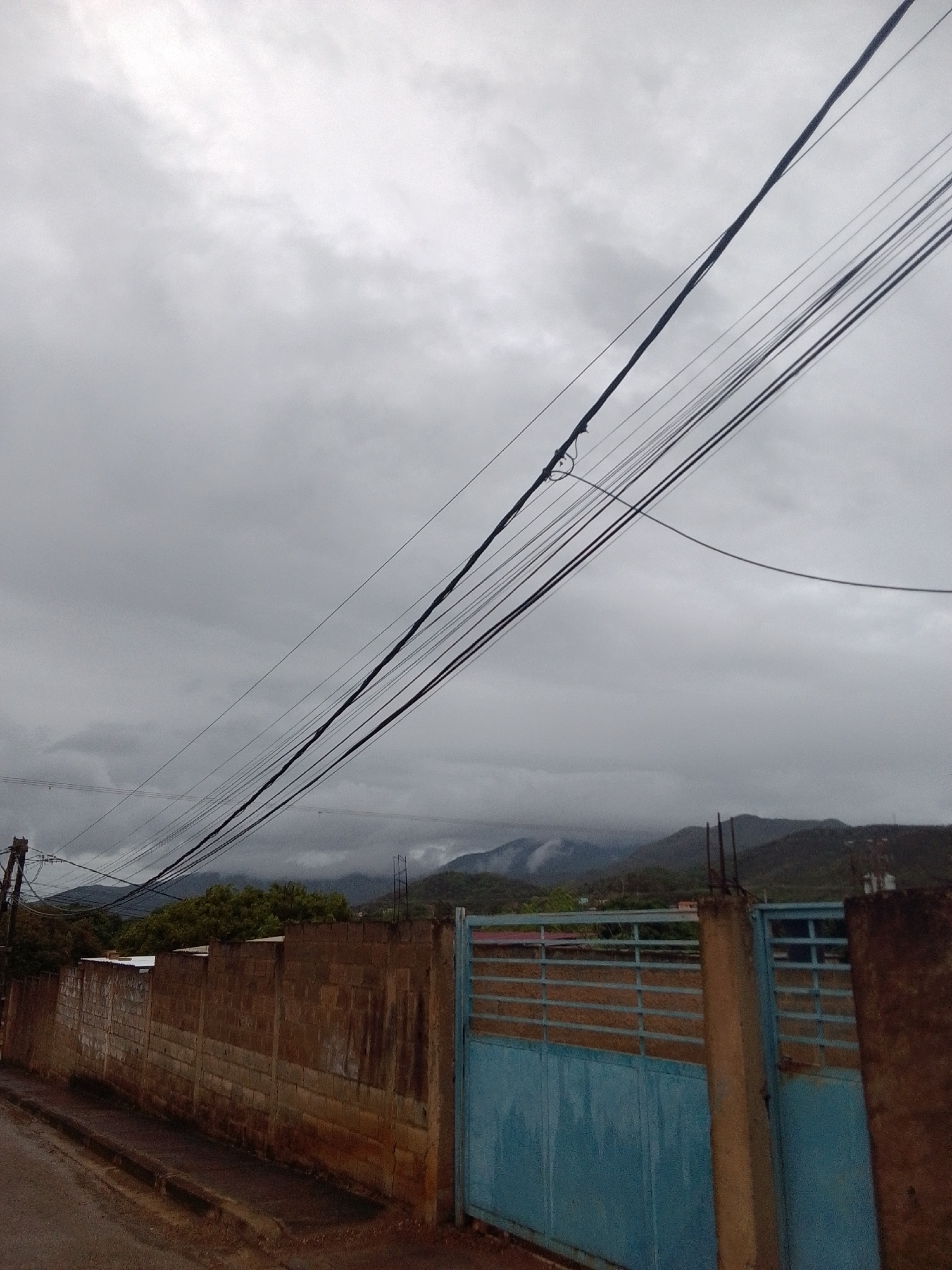 This image depicts an outdoor scene with a focus on utility wires stretched across a cloudy sky. In the foreground, a brown brick wall runs horizontally, with a blue metal gate visible on the right side. The wall appears weathered, with some sections showing signs of damage or age. The gate is constructed from horizontal metal bars. A dirt road or path is visible in front of the wall. In the background, rolling hills or mountains are shrouded in mist, suggesting a humid or overcast atmosphere. Dense green trees are situated between the wall and the hills. The sky is heavily overcast with gray clouds, indicating a day with poor visibility or impending rain. No people are visible in the image. The time of day is unclear due to the overcast conditions, but it appears to be daytime. The overall mood is subdued, possibly conveying a sense of a quiet or slightly melancholic atmosphere due to the weather and muted colors. There is no visible text in the image. The context of "La Tagua, Venezuela" suggests a rural or semi-rural mountainous region.