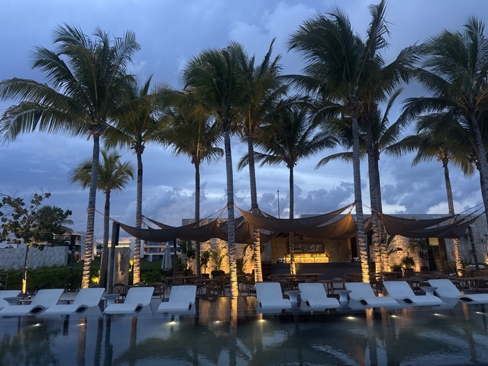 A tranquil scene unfolds with palm trees silhouetted against a cloudy twilight sky. Below, lounge chairs line the edge of a still pool, mirroring the illuminated structures and palm reflections. Dark canopies stretch between trees, hinting at a bar or outdoor dining area bathed in warm light. The atmosphere is peaceful and inviting, typical of a tropical resort at dusk.