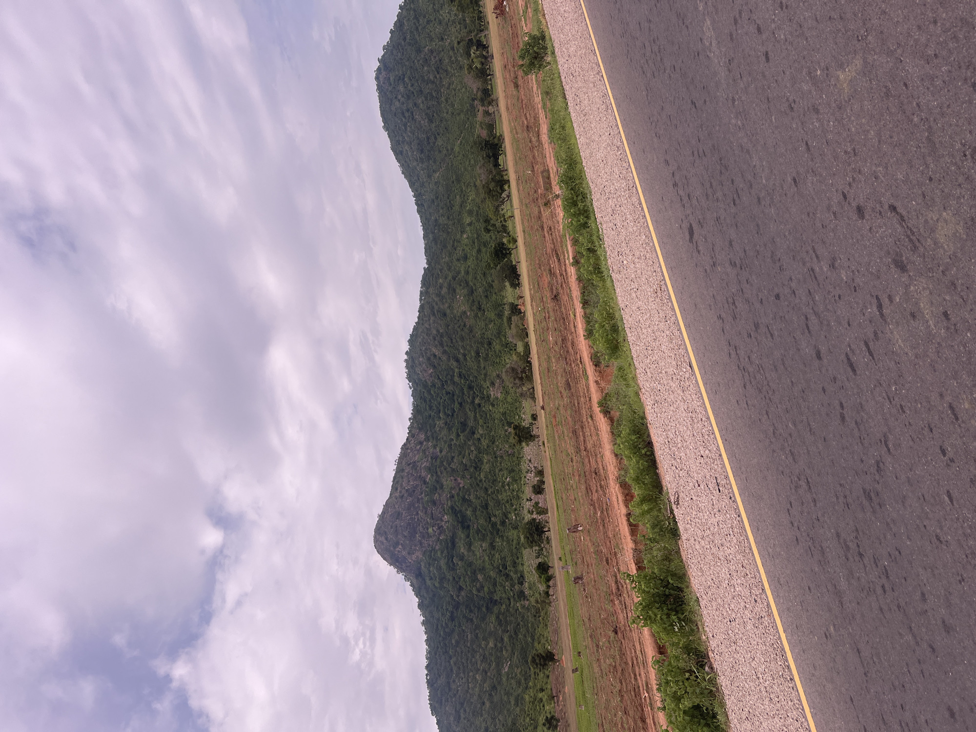 The image displays a roadside view with a prominent mountainous landscape on the left and a paved road on the right. The mountain is covered in dense green vegetation, suggesting a lush environment. A strip of lighter-colored ground, possibly dirt or gravel, with some sparse grass and a few scattered trees, separates the mountain from the road. The road itself is dark asphalt with a distinct yellow line running along its edge. The sky is partly cloudy with soft, diffused light, indicating it might be daytime, possibly morning or late afternoon. There are no people or discernible objects in the immediate foreground or on the road. The overall scene evokes a sense of natural beauty and a rural or semi-rural setting, likely in a location with hilly terrain and a road infrastructure. There is no visible text in the image. The orientation of the image suggests it was taken from a moving vehicle or a slightly tilted perspective.