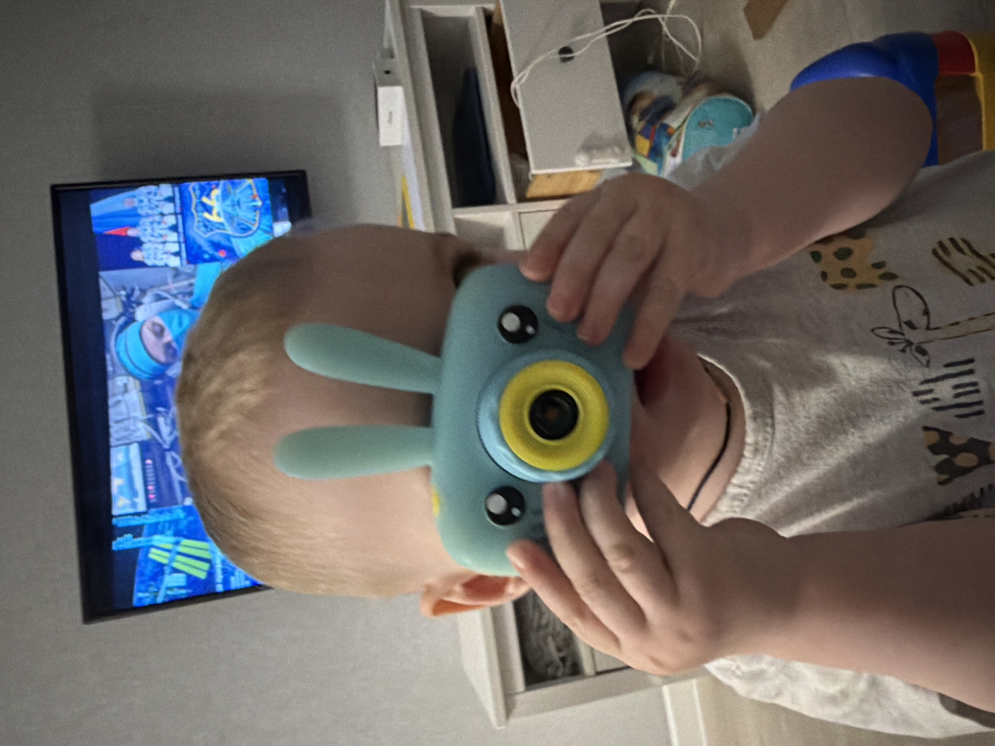 A child holds a toy camera with rabbit ears in front of his face. A television screen displays a video game. The background includes a cabinet and floor. It is not possible to confirm the location is Votkinsk, Russia.