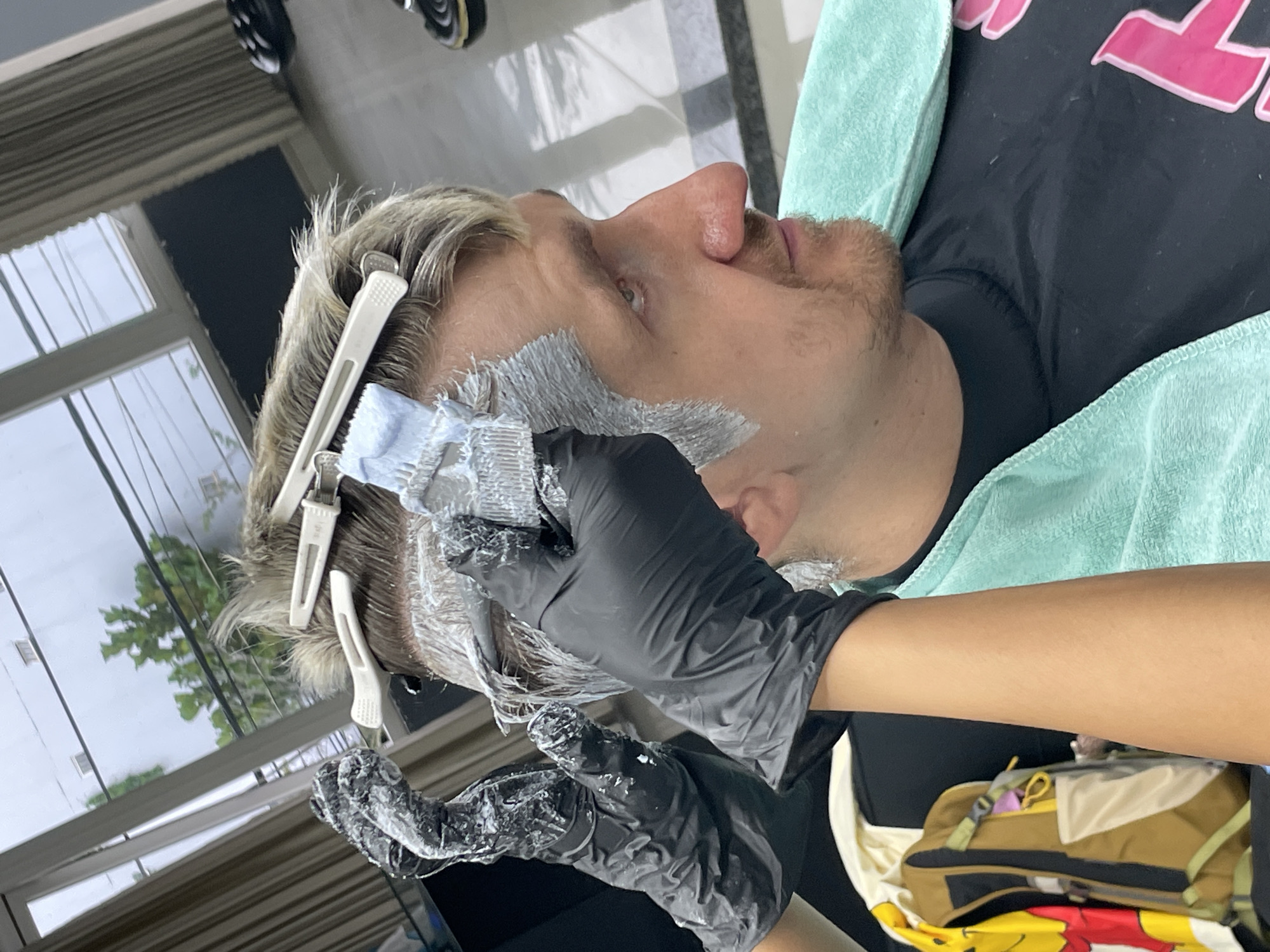 In an indoor hair salon located in Xuân Hương - Đà Lạt, Vietnam, a person is receiving a hair coloring treatment. Two gloved hands, one on the left and one on the right, are applying a light-colored substance to the individual's hair. The individual, a man with light-colored hair, is seated and looking to the right. He is wearing a black t-shirt and a light blue towel is draped around his neck. In the background, a window shows greenery and buildings, suggesting an urban environment. A backpack with a yellow character is visible to the left of the frame.