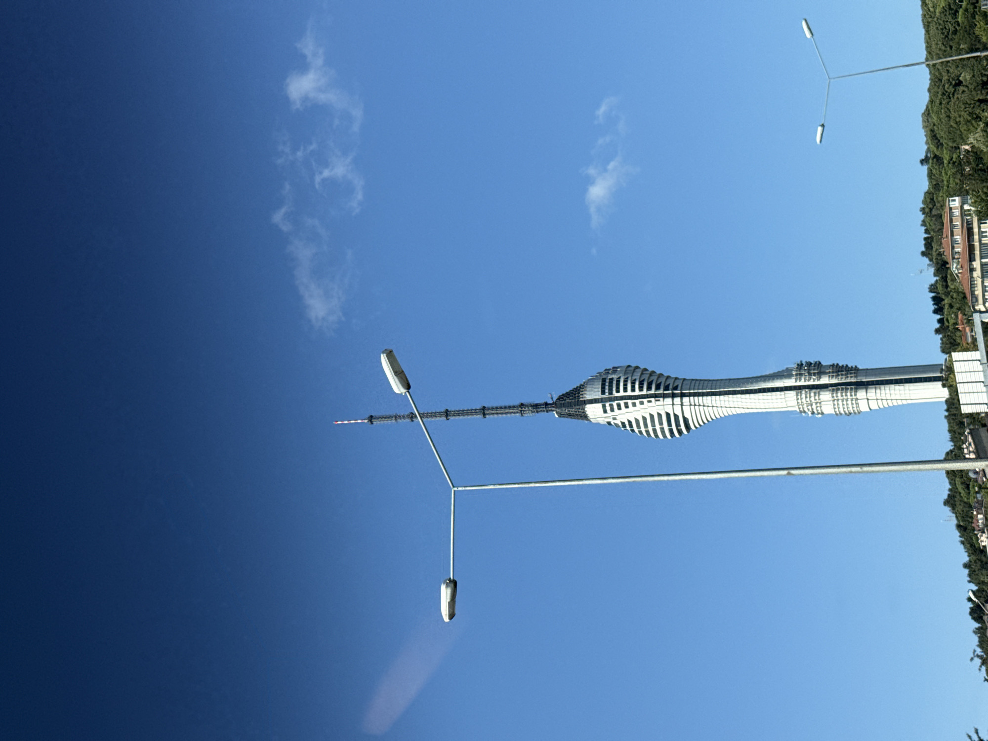 An outdoor, eye-level shot captures the Çamlıca Tower in Üsküdar, Turkey, against a clear blue sky. The tower, a prominent telecommunications tower with a distinctive twisting design, is the central subject. To the left, a modern street lamp with two lights stands in the foreground, its metallic pole extending upwards. Another street lamp is visible in the lower right corner of the image. The lower portion of the frame shows a tree-lined hill dotted with buildings and a few smaller structures. The scene appears to be captured on a bright, sunny day.