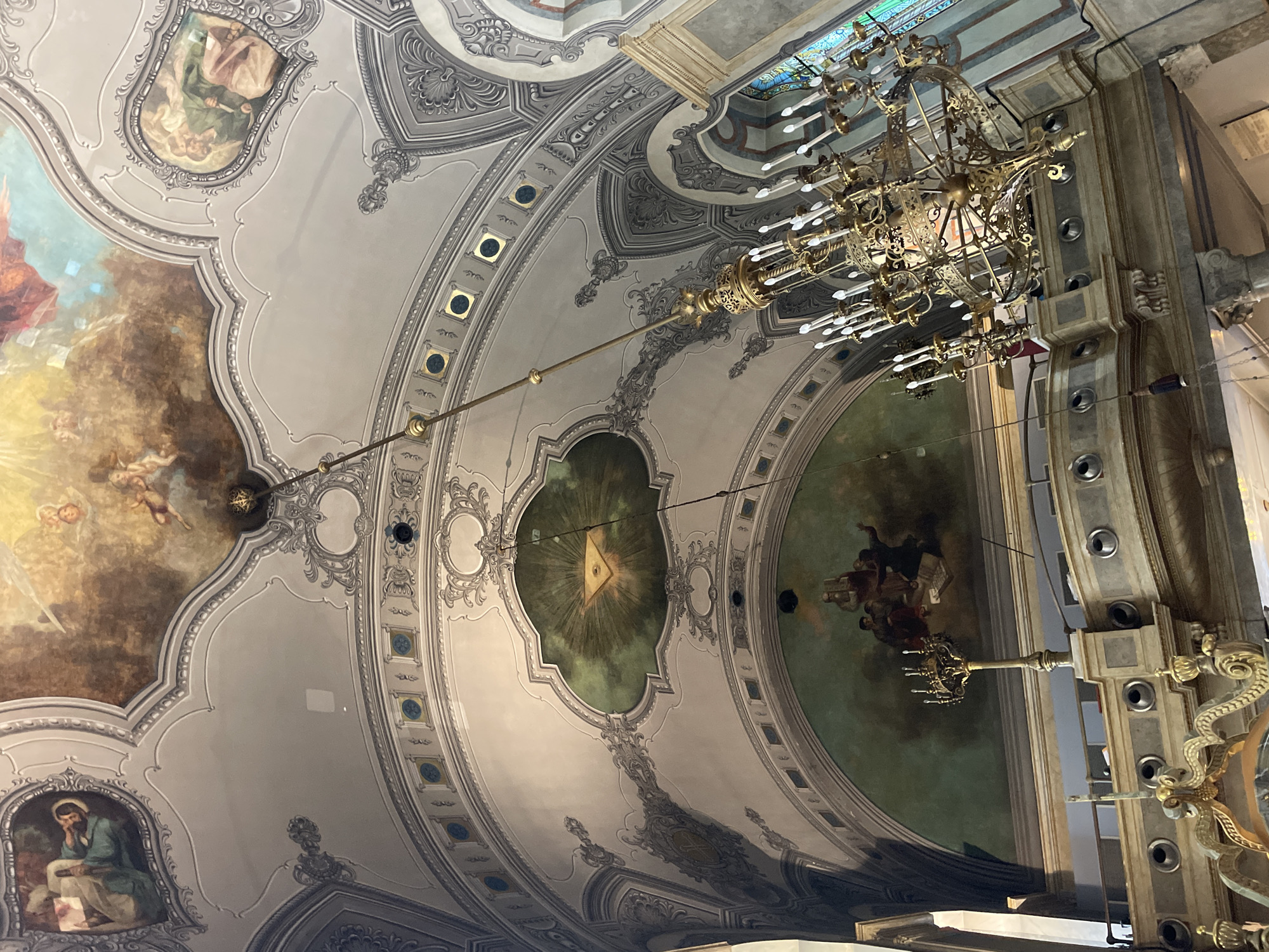 The image displays an upward view of a highly ornate ceiling, characteristic of a grand architectural structure, likely a church or cathedral in Novi Sad, Serbia. The ceiling is richly decorated with elaborate stucco work, featuring intricate patterns, moldings, and framed sections in shades of grey and white, accented with gold.
A large, multi-tiered brass or gold chandelier with numerous electric candle-shaped bulbs hangs prominently from the ceiling, dominating the right side of the frame. The ceiling design incorporates several large paintings and smaller medallions.
A notable painting in a central, green-hued medallion depicts the Eye of Providence – a golden eye within a triangle, surrounded by rays of light. Other paintings show religious or allegorical figures: one depicts a bearded man, possibly a saint or evangelist, seated and writing on a scroll, while another section illustrates celestial figures, including angels or cherubs, bathed in a warm, glowing light. A third painting shows a group of figures in an indeterminate scene.
The overall style is elaborate, suggesting Baroque or Rococo influences, with an emphasis on detailed craftsmanship and narrative art. The combination of religious iconography, grand-scale paintings, and ornate architectural elements indicates a significant place of worship or public building. The lighting is primarily artificial from the chandelier, with hints of natural light through what appear to be stained-glass windows in the upper right. No legible text is visible in the image.