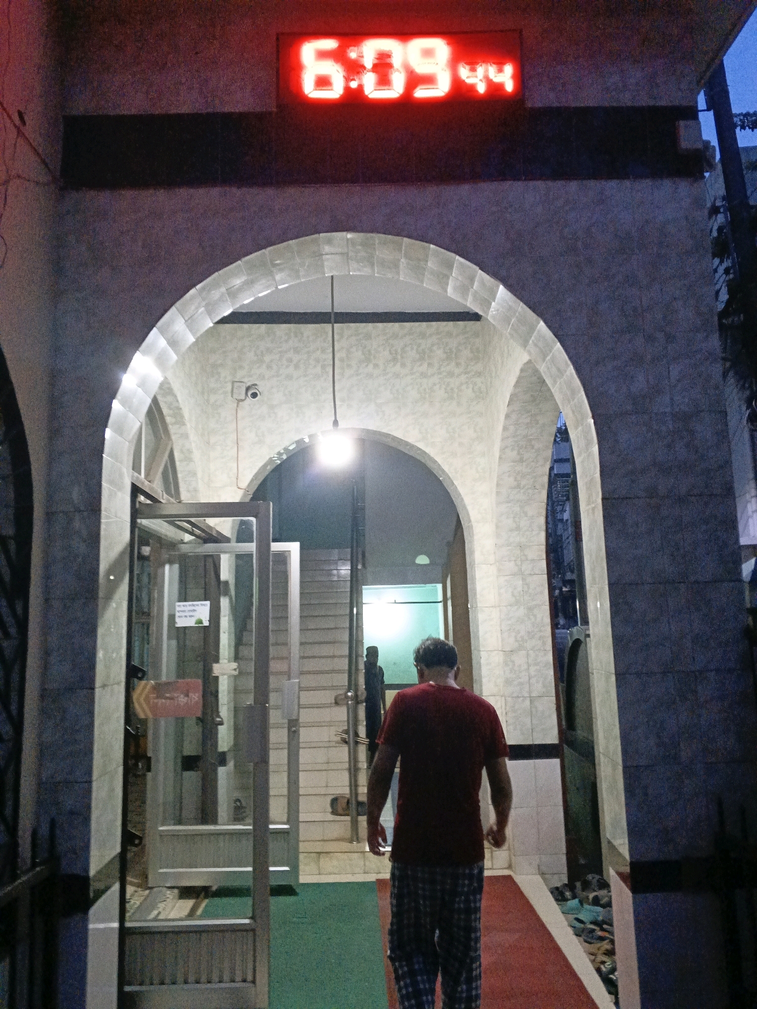 An overhead red digital clock displays "6:09". A man wearing a maroon t-shirt and plaid shorts walks away from the camera through an archway. To his left, glass doors with metal frames lead into a room. The floor inside the archway is a combination of red and green carpet with white trim. The overall setting appears to be the entrance to a building, possibly a mosque, in Sylhet, Bangladesh.