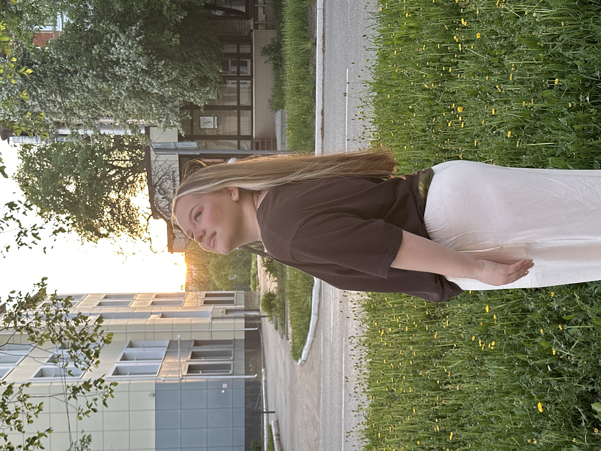 A young woman stands outdoors on a warm, sunny day. She has long, light brown hair with blonde highlights, and is wearing a dark brown short-sleeved shirt and white pants. She is looking towards the sun, and her arms are relaxed at her side.
The location is Chaikovsky, Russia. The woman is standing next to a grassy area filled with dandelions. A sidewalk runs alongside the grass, and a street and modern multi-story building are visible in the background. Trees are also visible, casting shadows on the scene. The setting sun provides a warm, golden light that illuminates the sky and the woman's face.