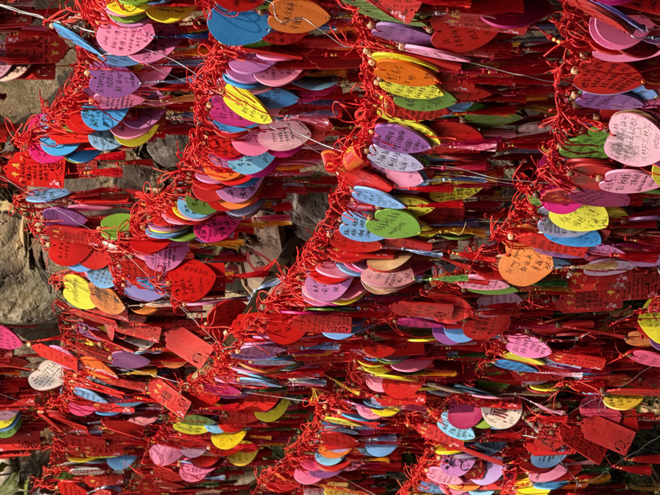 A dense array of colorful heart-shaped and rectangular wooden plaques hangs by red knotted strings. The plaques are densely packed and cover a stone wall. Colors include red, pink, blue, yellow, orange, green, and purple. Many plaques are covered in writing. The overall impression is a vibrant and intricate tapestry of wishes or messages. Sunlight shines on the scene, enhancing the colors.