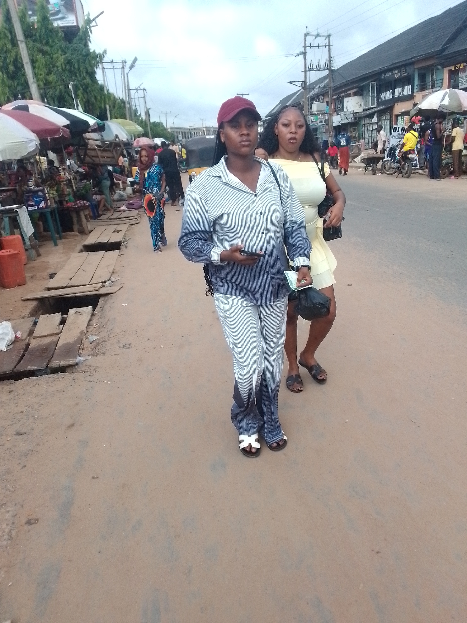 Two women are walking down a dirt road in Egbagu, Nigeria. The woman in front is wearing a striped pantsuit and a maroon baseball cap, and is holding a phone. The woman behind her is wearing a short yellow dress. The background shows a bustling street scene with shops, people, and vehicles. There are wooden boards covering some of the drainage ditches on the side of the road. There are market umbrellas on the left side of the frame, indicating a marketplace is nearby. Powerlines are visible in the background. The ground is a dirt road with signs of wear.