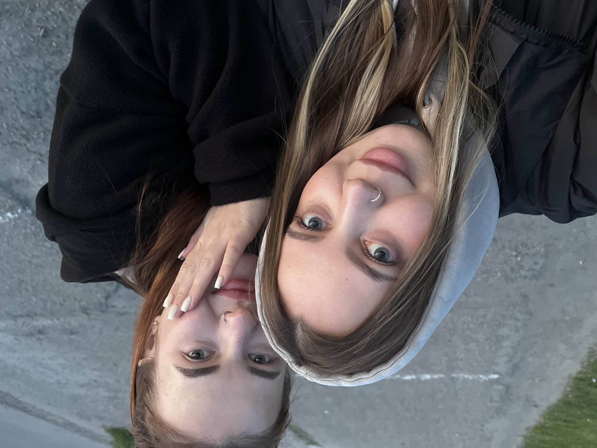 Two young women are featured in a close-up, rotated outdoor photograph taken in Novopokrovskaya, Russia. The woman on the right (as viewed upright) has long, brown hair with lighter highlights, blue eyes, and a silver nose ring. She wears a grey hooded top layered under a dark, puffer-style jacket. Her lips are a light pink, and she has visible eyeshadow. The second woman, positioned to the left, has reddish-brown hair and green eyes. She wears a black long-sleeved top or sweatshirt. Her right hand, featuring long, light-colored fingernails, is gently placed near the other woman's face. Both women look directly at the camera with neutral to slightly pleasant expressions, suggesting they are posing for a selfie or a close-up portrait together. The background is a soft blur of grey, textured ground, likely asphalt or concrete, with a patch of green grass visible in one corner. The lighting appears natural and soft, indicative of daytime, possibly overcast conditions, and the jackets worn suggest mild weather. There is no visible text in the image.