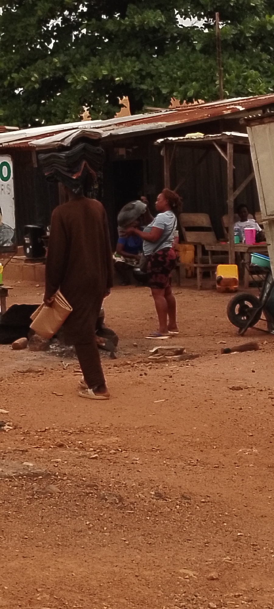 This image depicts a street scene in Gboko, Nigeria. Two individuals are visible. In the foreground, a person is walking away from the camera, carrying a stack of folded items on their head and a paper bag in their right hand. This person is wearing a brown garment and white sandals. To their right, a woman is standing and holding a large, dark circular object in her arms. She is wearing a light blue t-shirt and a patterned skirt, and she is also wearing sandals. Behind them, other people are seated at tables, and various objects, including containers and a wheelbarrow, are scattered around the area. The environment appears to be outdoors, with dirt ground and a rustic structure in the background, suggesting a market or informal gathering space.