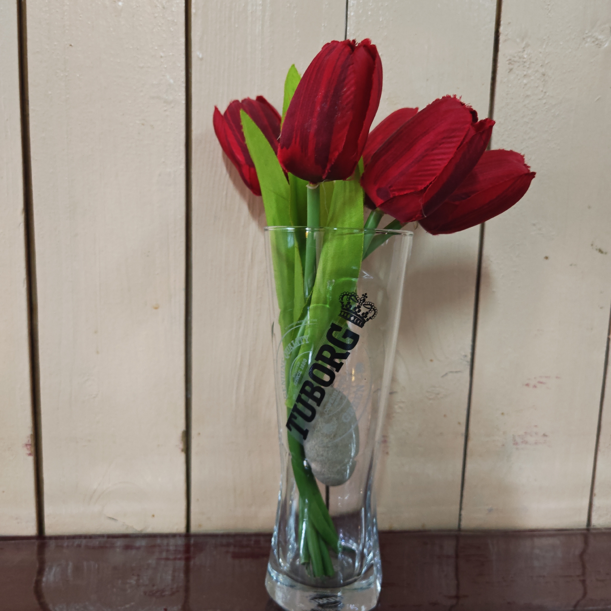 A glass vase with the "Tuborg" beer brand logo is positioned on a dark brown surface, presumably a table. The vase contains a bouquet of red tulips with green stems and leaves. The background is a light-colored wall with vertical panels. The location is unknown.