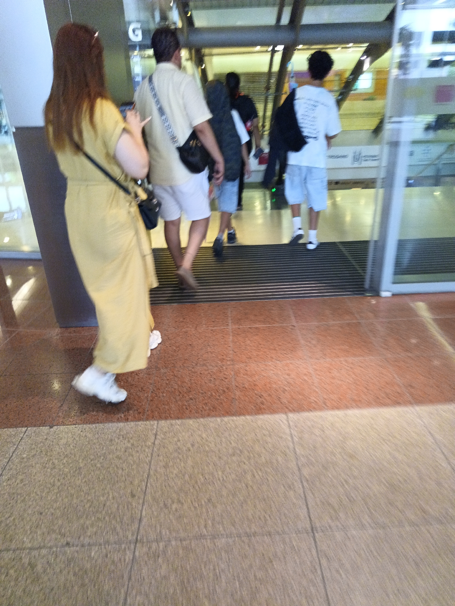 The image depicts a scene inside an air-conditioned environment, likely an airport terminal or a shopping mall, with a polished orange and grey tiled floor. Several individuals are visible. On the left, a woman with long brown hair is walking away from the camera, wearing a yellow dress and white sneakers. She is carrying a black purse over her shoulder. To her right, a man in a light-colored shirt and white shorts walks ahead, also with a black bag. Further ahead, a person in a white t-shirt and light blue shorts is walking with a black bag slung over their shoulder. Another person, partially visible on the far right, appears to be in blue shorts and white socks. The overall lighting is bright, and the environment suggests a public transit hub or commercial space. The presence of glass doors and industrial-style overhead structures further supports this interpretation. This scene could be located in Kuala Lumpur, Malaysia, a major city known for its modern infrastructure and bustling transportation networks.