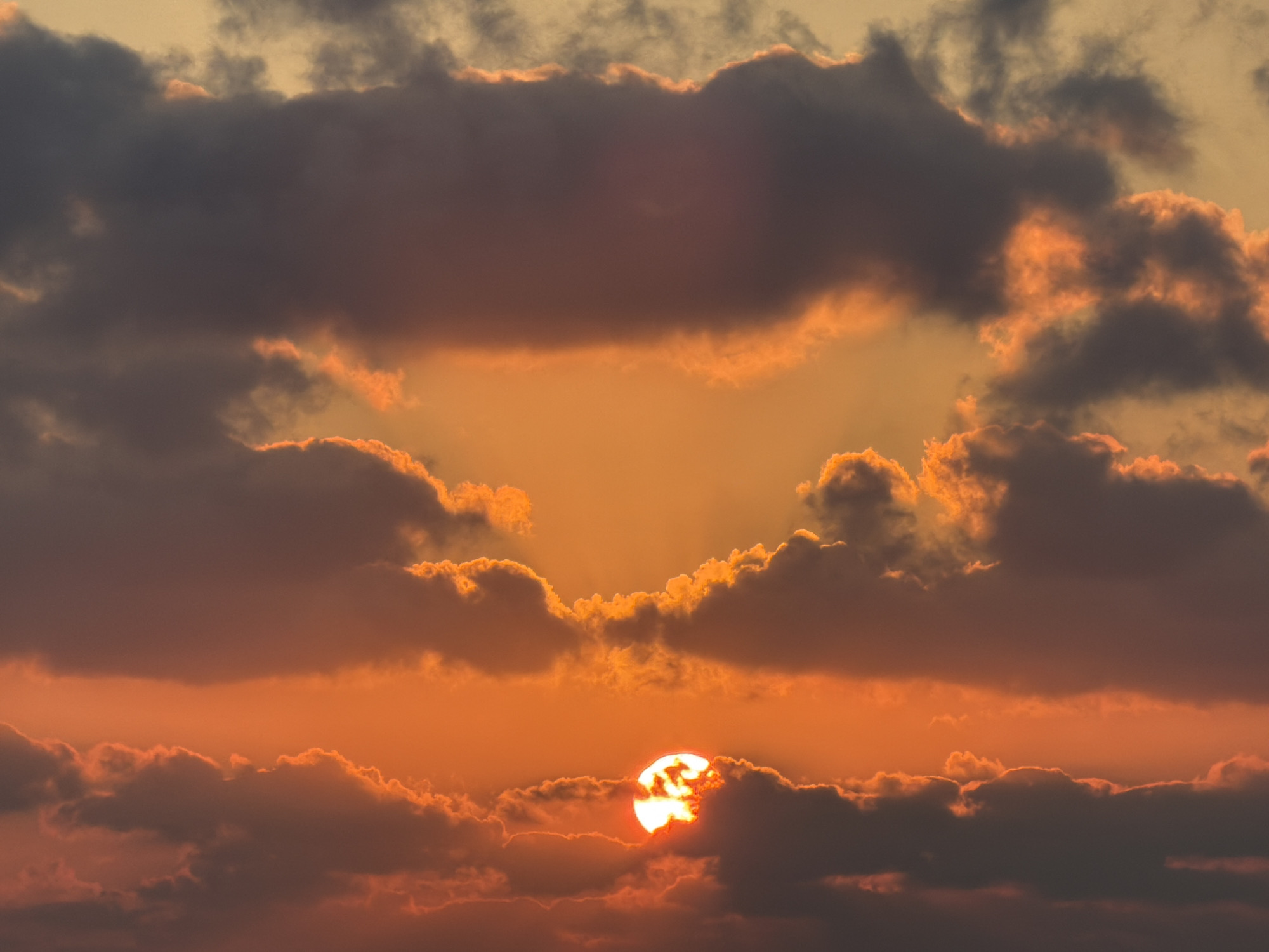 The image depicts a dramatic sunset over the city of Tel Aviv, Israel. The sun, a bright, glowing orb, is partially obscured by dark, voluminous clouds that are illuminated with fiery orange and pink hues. These clouds create a striking contrast against the warmer, golden sky. The overall scene is one of intense natural beauty, with the clouds forming an abstract, almost heart-like shape around the radiant sun. No people or specific landmarks are visible, focusing solely on the atmospheric phenomenon.