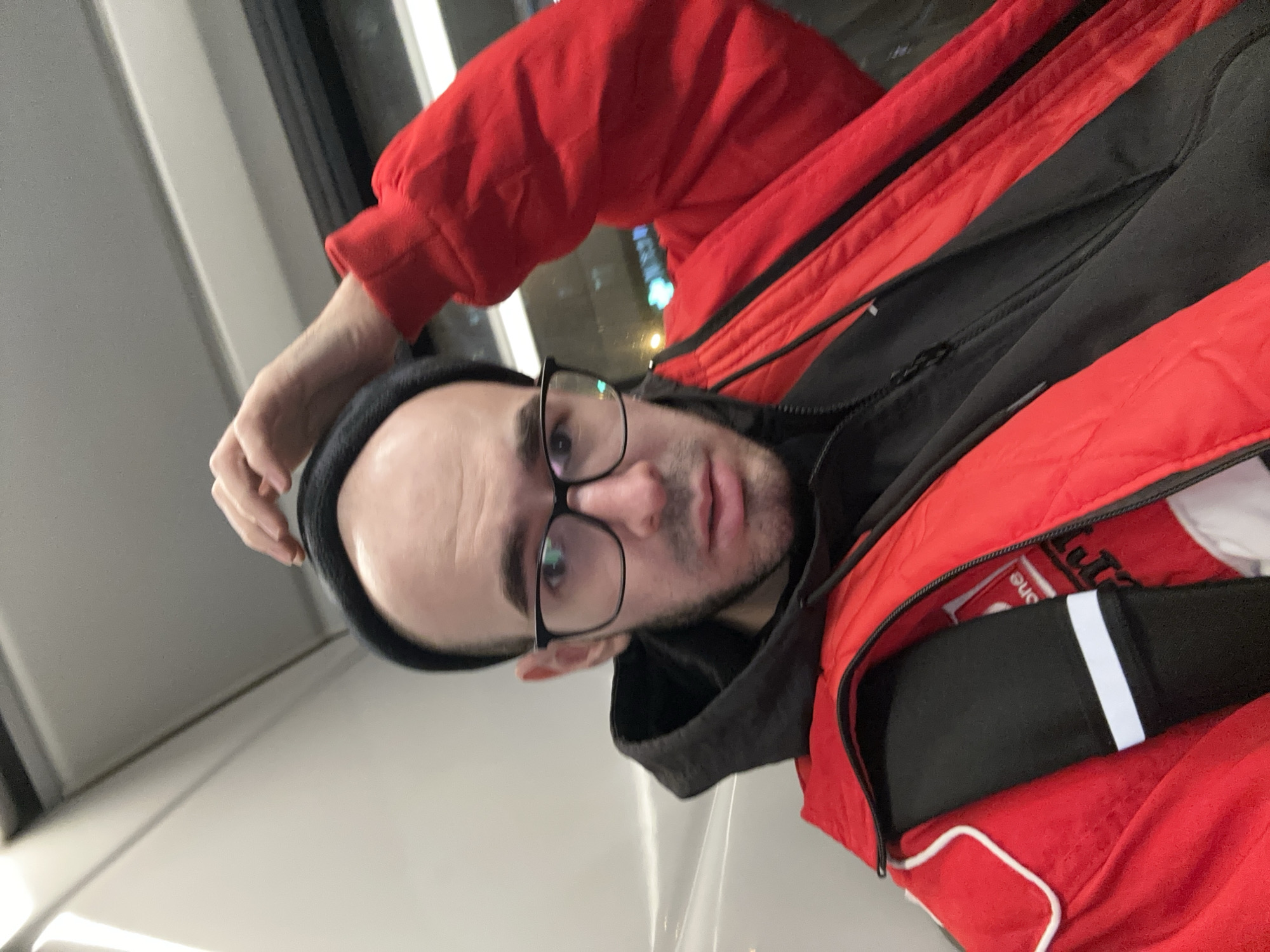 A man wearing glasses and a black beanie, positioned on what appears to be public transportation, possibly a bus or train, is the central subject. He is wearing a red and black jacket. It's dusk or nighttime as evidenced by the reflection of streetlights or city lights visible through the window behind him. There is no discernible evidence within the image to confirm its location within the city of Khimki, Russia.