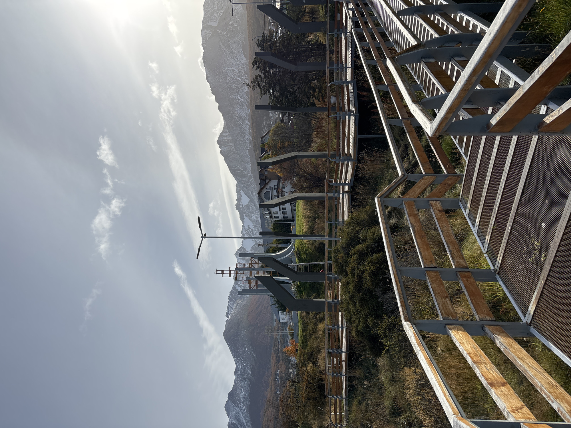 The image shows a pedestrian walkway or bridge in Ushuaia, Argentina, with snow-capped mountains in the background. The walkway is constructed of metal and wood. The sky is partly cloudy. A street light is visible near the walkway. A building is partially visible amongst the trees.