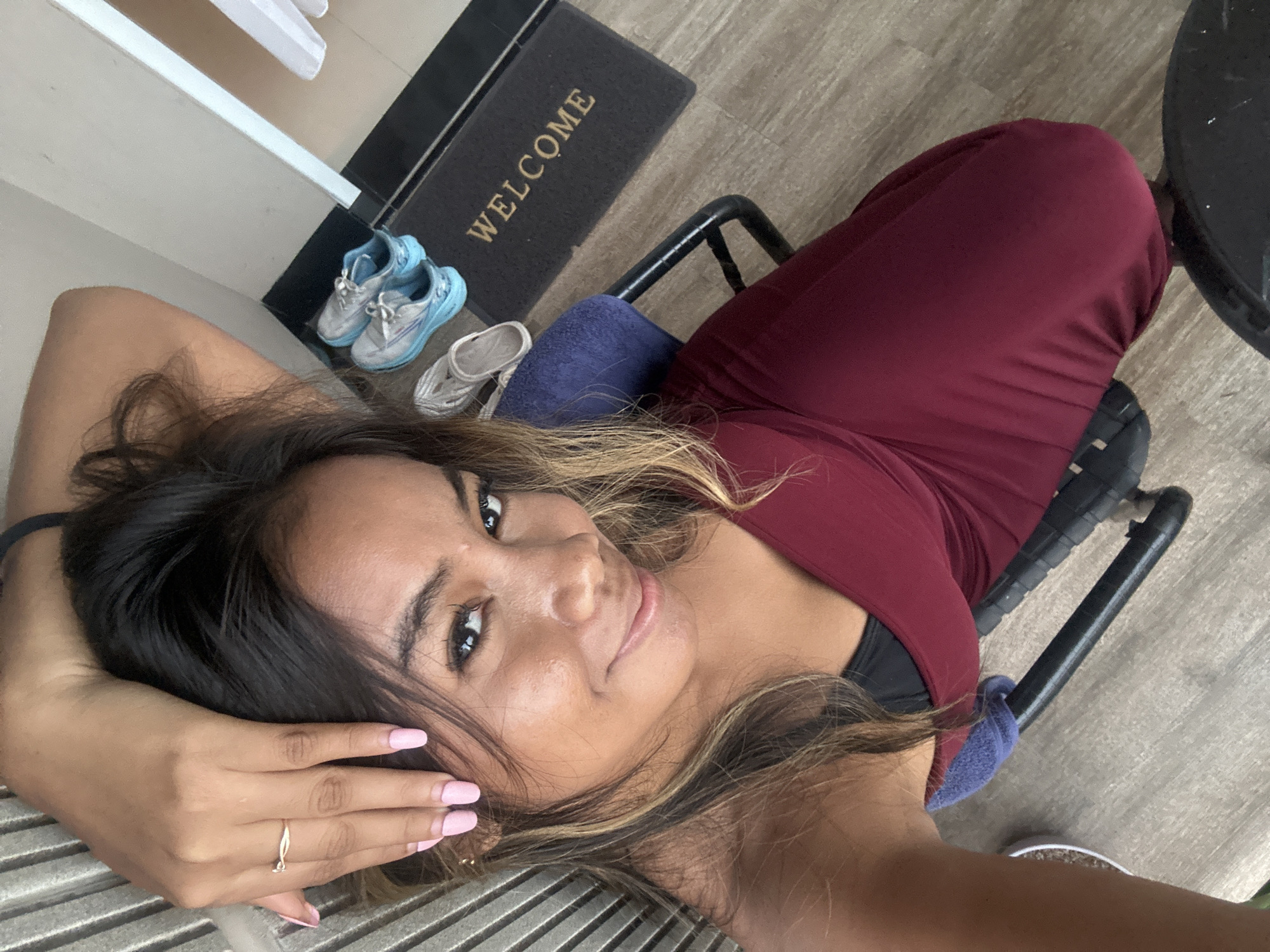 The image shows a woman lying down with her head resting on her hand. She is wearing a maroon dress and a black top underneath. Her fingernails are painted pink. In the background, there is a welcome mat and a pair of blue and silver shoes. The flooring appears to be light-colored wood. The scene suggests an indoor environment, potentially a hotel room or a private residence. The presence of a welcome mat and the casual pose of the woman could indicate she is at a hospitality establishment in Kuta, Indonesia, a popular tourist destination known for its beaches and nightlife.