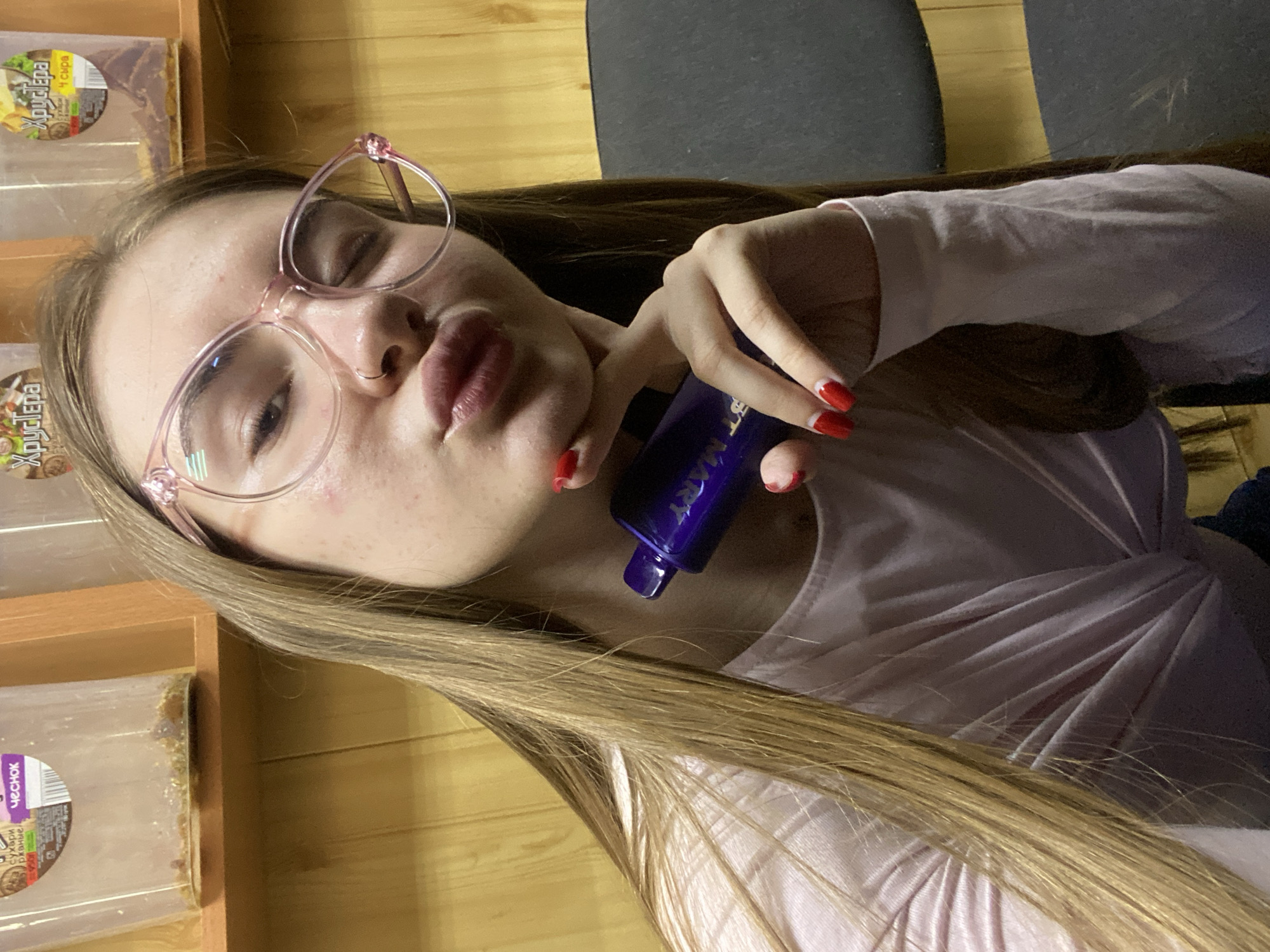 A young woman with long blonde hair and pink-rimmed glasses poses for a selfie, making a pouty face. She is wearing a light pink long-sleeved shirt. Her nails are painted red. She holds a small, dark blue bottle with some text on it in her right hand. The background appears to be a wooden wall, with shelves holding various food items, some labeled with Russian text. One label visible in the top left corner reads "Хрустёпа 4 сыра," which translates to "Khryustyopa 4 cheeses." Another label on the lower left reads "Чеснок," meaning "Garlic." The lighting suggests an indoor setting, possibly a kitchen or pantry. The overall mood is playful and casual.
