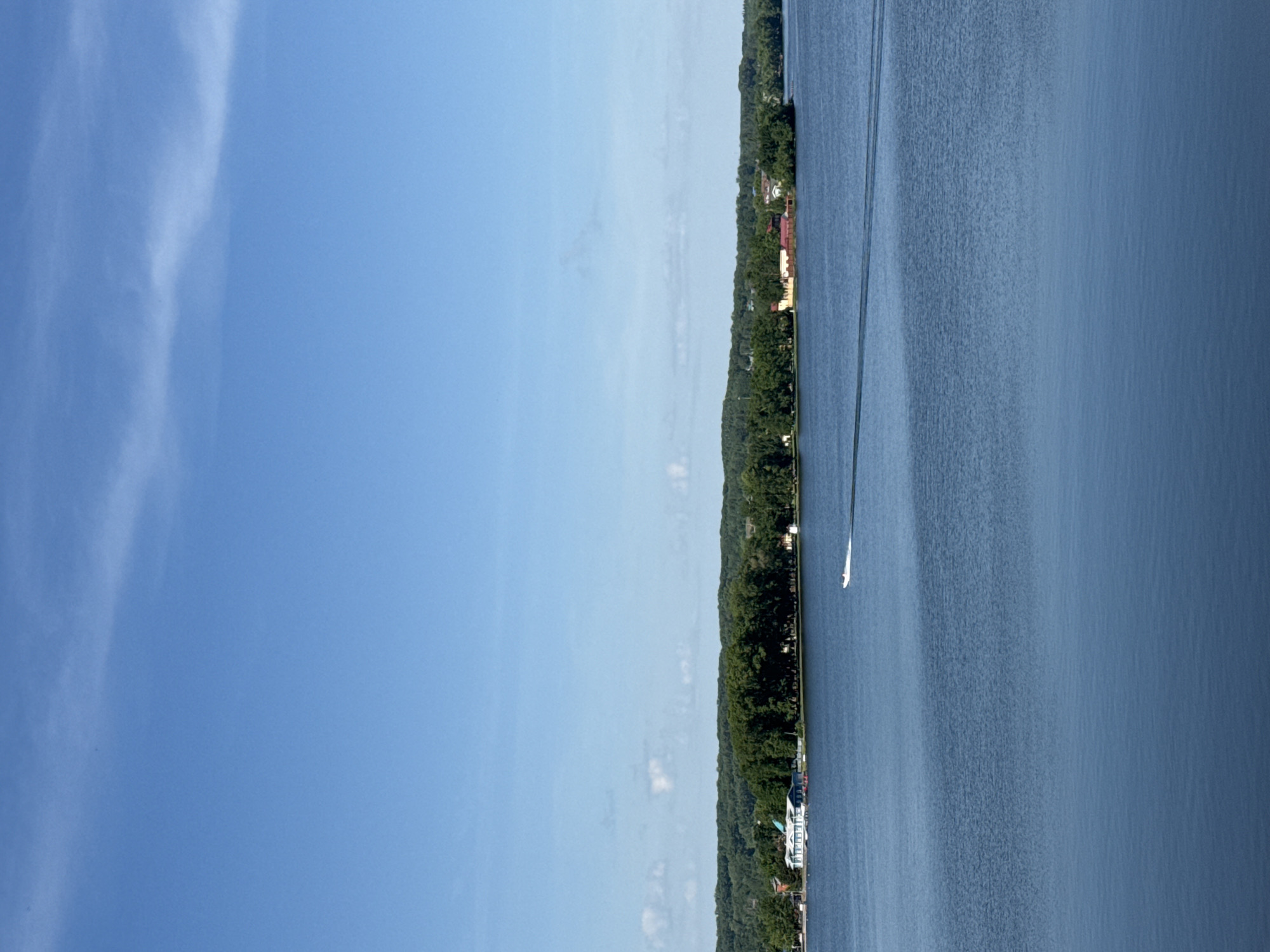 The image shows a water landscape, likely a lake or wide river, with a forested shoreline on the right side. A boat is moving on the water, leaving a visible wake. Some buildings, including what appear to be houses, are visible along the shoreline amidst the trees. The left side of the image features a clear blue sky with a few wispy clouds. Without other information, it is not possible to confirm if the photo was taken in the city of Votkinsk, Russia.