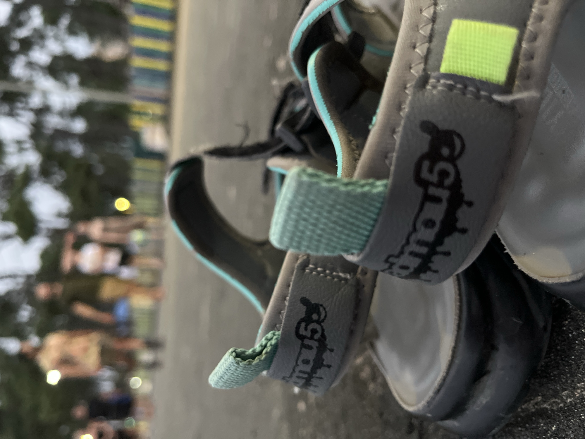 The image is a close-up, low-angle shot of a pair of grey and teal sandals with a black sole. The sandals are positioned on a dark, textured surface, likely asphalt or concrete. On the side of one sandal, a logo is visible, featuring the text "mou5" in black with a dripping effect, accompanied by a cartoonish face with circular eyes and a curved mouth. A small, lime green square tag is attached to the same sandal strap. In the background, the scene is out of focus, showing several blurred figures of people walking. Behind them, a blurred line of trees and a yellow and blue striped barrier are visible. The lighting suggests it is late afternoon or early evening, with some artificial lights glowing in the distance. The image appears to have been taken outdoors, possibly in a public area such as a park or promenade, consistent with a location in Ureki, Georgia, a resort town known for its beaches and walking paths.