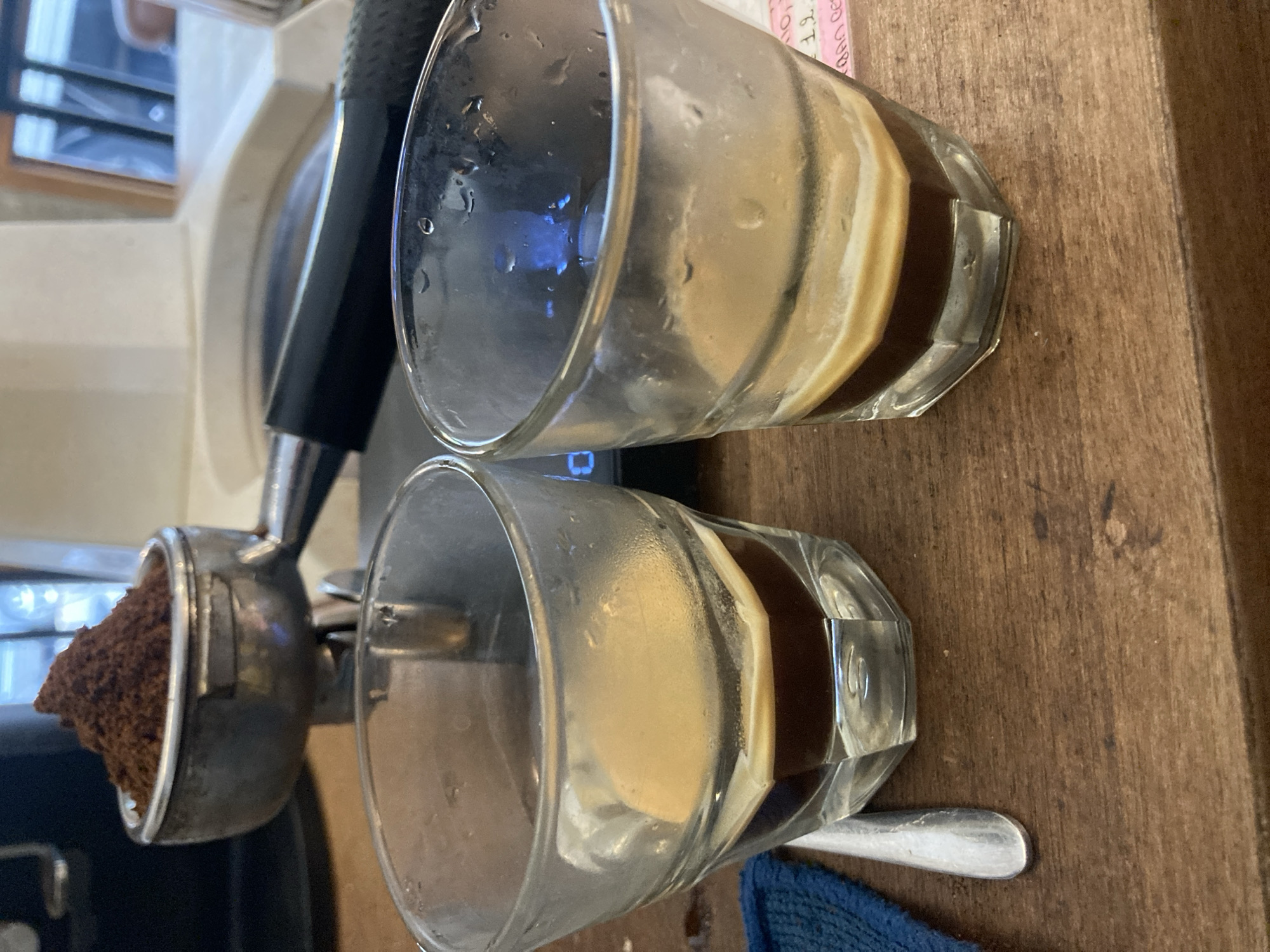 The image shows a close-up of an espresso machine and two glasses of coffee. The espresso machine is on the left side of the frame, with a portafilter filled with ground coffee. The glasses of coffee are on the right, sitting on a wooden surface. They appear to be layered with a dark liquid (likely espresso) at the bottom and a lighter, creamy liquid (likely milk or cream) on top. The glasses are octagonal in shape. A teaspoon rests near one of the glasses, and a blue cloth is partially visible. The background is blurred, suggesting a shallow depth of field. The location is unknown.