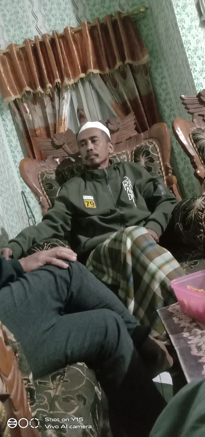 The scene is set indoors in Gading Kulon, Indonesia. A male individual is seated on an ornate wooden armchair with dark patterned upholstery. He wears a white knitted skullcap, a dark olive green zip-up jacket displaying a yellow "79" patch on the left chest and white text on the right, and a plaid sarong in shades of green, brown, and white. His left arm rests on the armrest, and his right hand is positioned on his lap. He is looking towards the left side of the frame. In the foreground, to the left of the primary subject, the lower torso and legs of another individual are visible, clad in dark trousers; a hand rests on their knee. The background features a wall covered with light green patterned wallpaper. A window is partially visible, dressed with sheer white curtains and an outer layer of shimmering reddish-brown fabric curtains with gold fringe, suspended from a gold-colored rod. A second, similar ornate wooden armchair is partially visible to the right. A pink rectangular plastic container rests on a dark patterned surface in the bottom right.