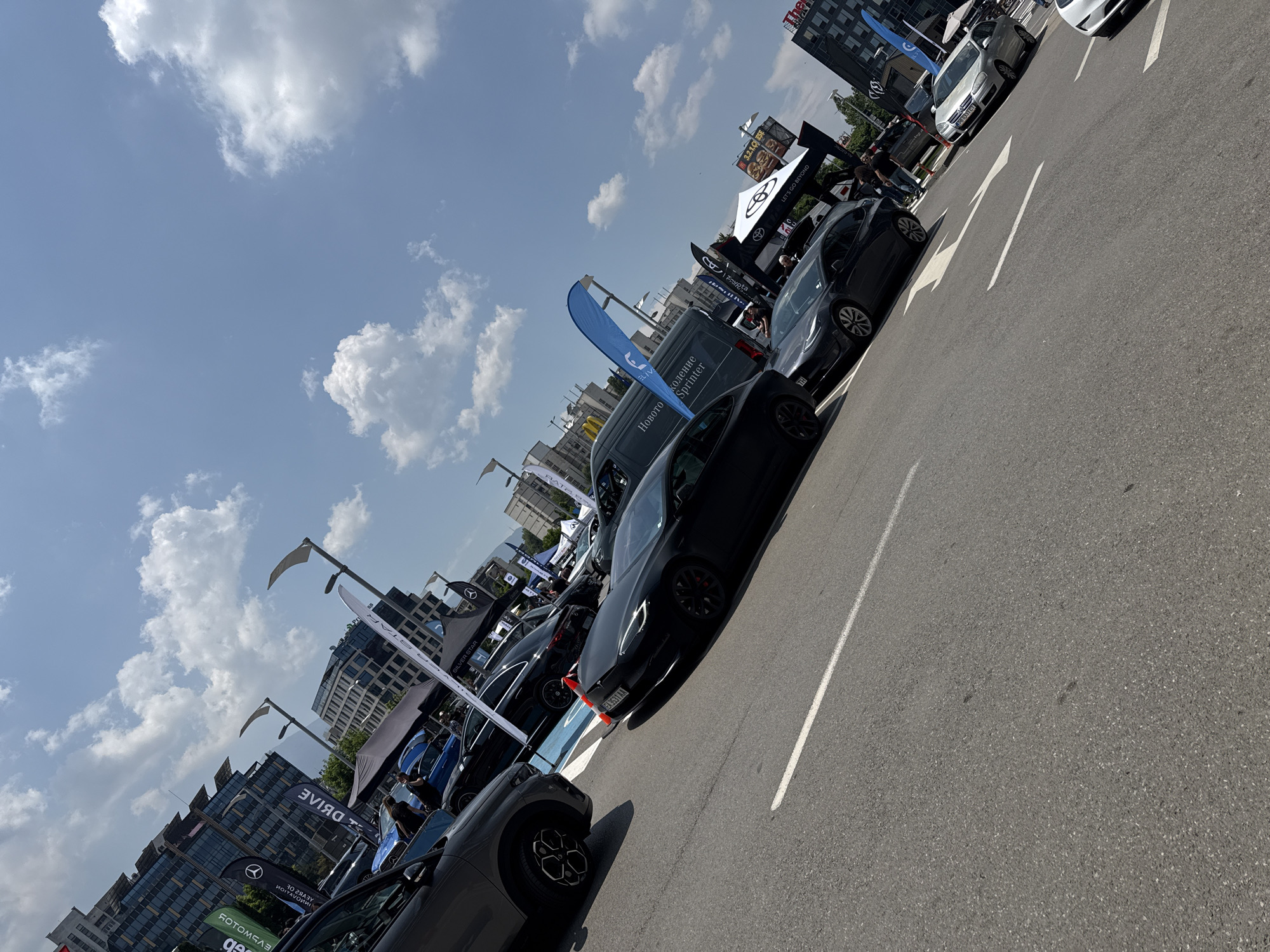 This image captures an outdoor car exhibition or sales event taking place on an asphalt surface, likely a large parking lot or street, under a clear blue sky with scattered white cumulus clouds. The scene is located in Sofia, Bulgaria, identified by the "CA" prefix on visible vehicle license plates (e.g., "CA 5143 AA" on a black Tesla), Bulgarian text on banners ("Новото поколение Sprinter"), and background elements such as a partial "The Mall" sign and McDonald's golden arches, consistent with the Tsarigradsko Shose Blvd. area.
In the immediate foreground on the left, the front quarter of a dark grey SUV with distinctive black and silver multi-spoke wheels is visible. A solid white line runs diagonally across the asphalt in the foreground. Further back, a black Tesla Model 3 or S, featuring red brake calipers, is parked diagonally across a section of the pavement painted blue, with an orange traffic cone positioned near its front left wheel. Behind it, another dark sedan, possibly a Mercedes-Benz, is parked.
Multiple promotional banners and tents are erected throughout the exhibition area. These include vertical banners with the Mercedes-Benz logo, "SILVER STAR," and reversed "eMotors" text, as well as a large blue teardrop-shaped banner. A black tent with "TOYOTA LET'S GO BEYOND" is visible in the mid-ground. A dark grey Mercedes-Benz Sprinter van is parked beneath one of the banners. Further down the asphalt, a silver Volkswagen Golf is visible, displaying another "CA" prefixed license plate.
Several individuals are present, distributed among the cars and tents, observing the vehicles and conversing, indicating their participation as attendees or staff at the event. The background features modern, multi-story buildings with glass facades; some buildings exhibit rooftop structures, possibly solar panels. Modern, curved streetlights are also integrated into the urban setting. The overall lighting suggests a sunny daytime environment.
